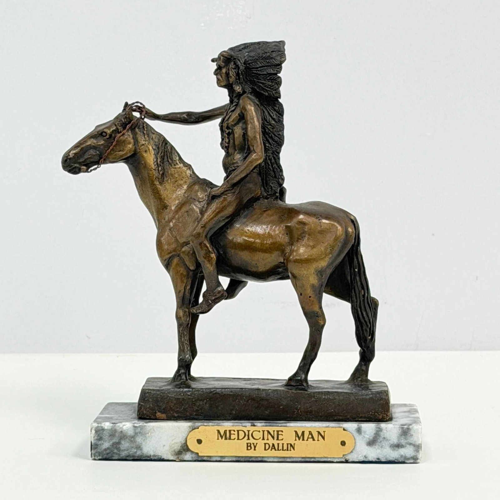 After Cyrus Edwin Dallin (American, 1861-1944) Bronze Indian Sculpture, Medicine Man, Signed: A Cyrus Edwin Dallin bronze sculpture titled Medicine Man that depicts a Native American Indian figure wearing an elaborate, full-feathered headdress seated on a horse. The figure holds a pair of thin