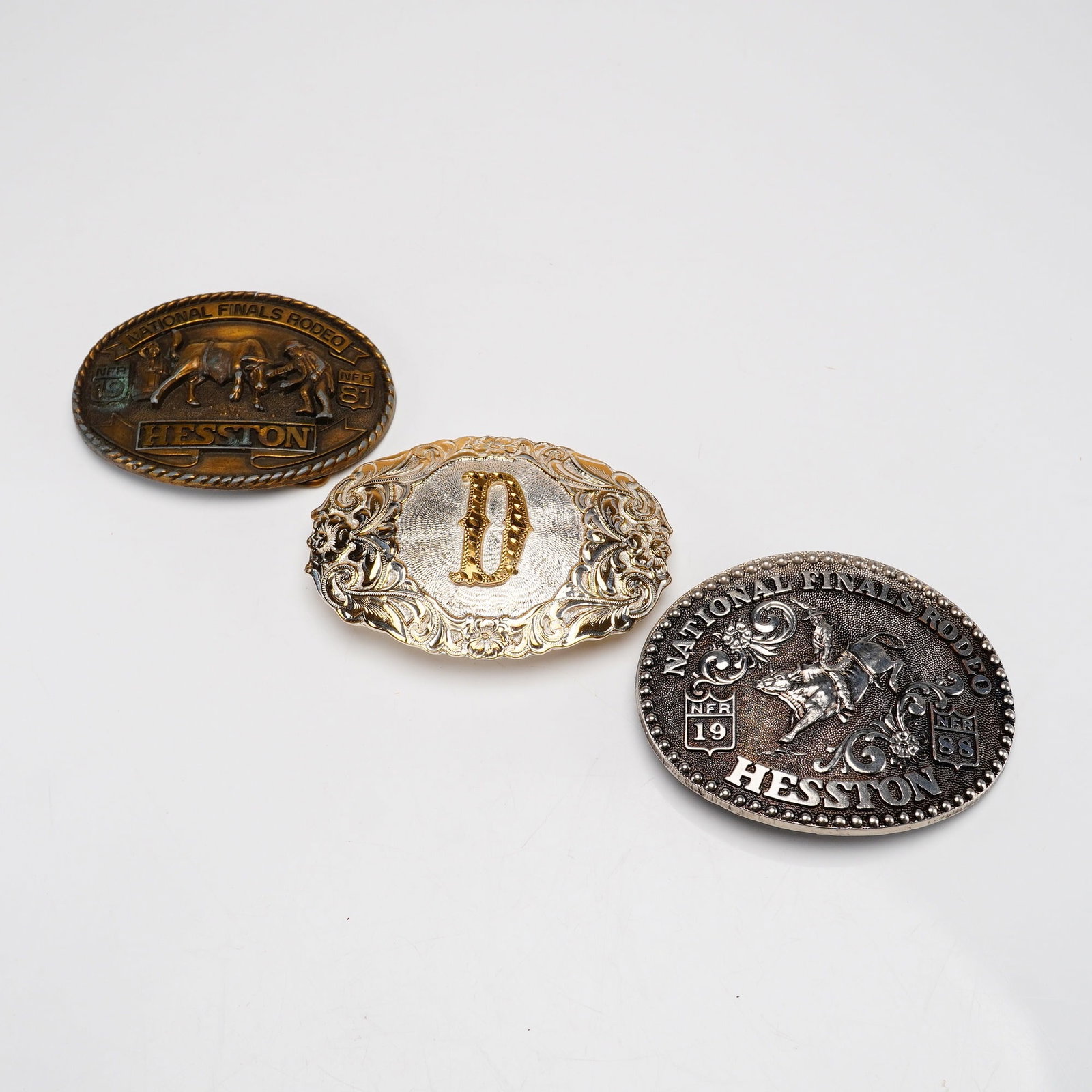 3pc Fred Fellows and Crumrine Metal Belt Buckles featuring Hesston: This lot of 3pc metal belt buckles includes two commemorative National Finals Rodeo (NFR) pieces and one monogrammed western buckle. The first item is a brass-toned metal buckle from 1981, identified 