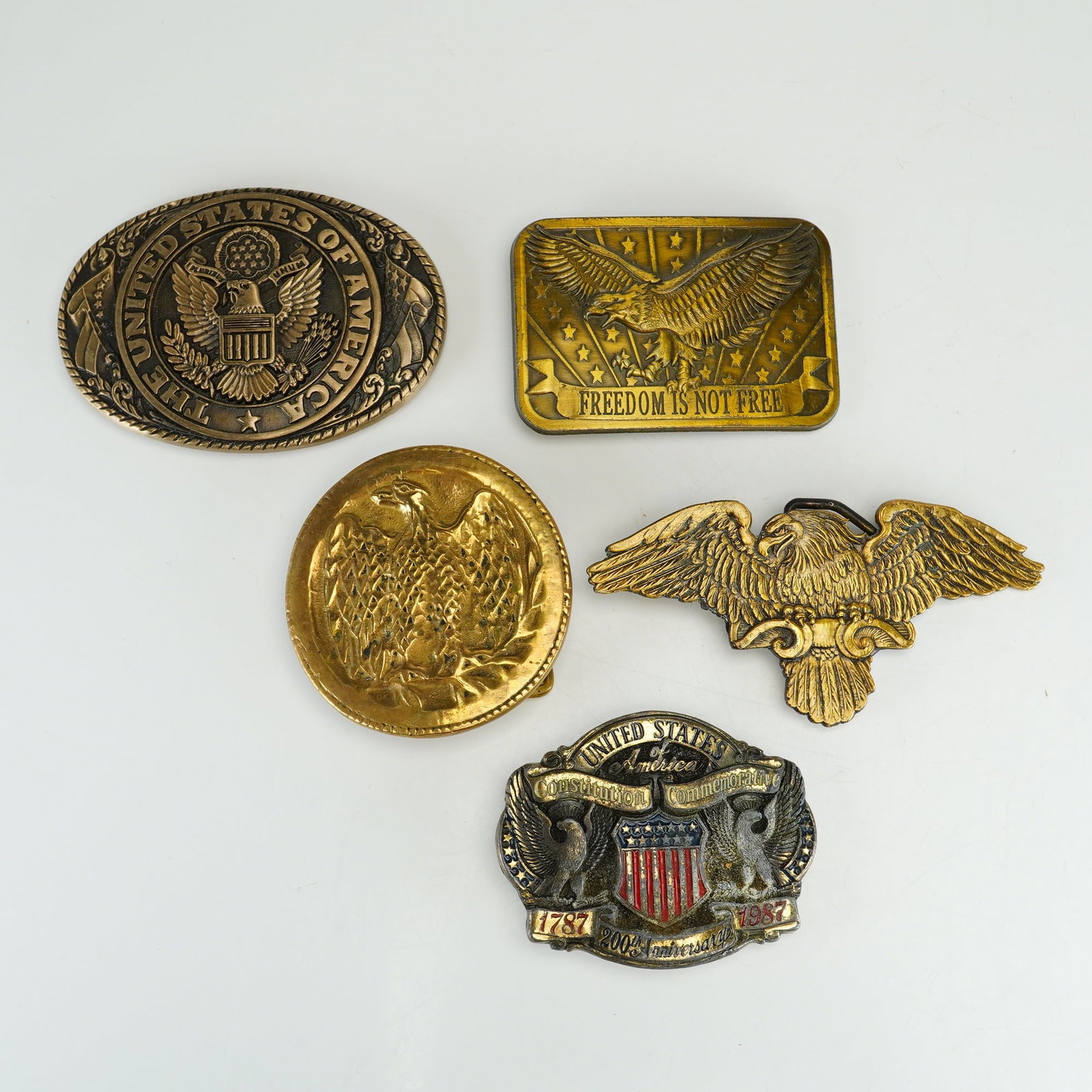 A Collection of Five Patriotic American Motif Metal Belt Buckles: This collection consists of five metal belt buckles featuring various American patriotic motifs and eagle designs. The first item is an oval solid brass buckle by Tony Lama featuring a raised relief o