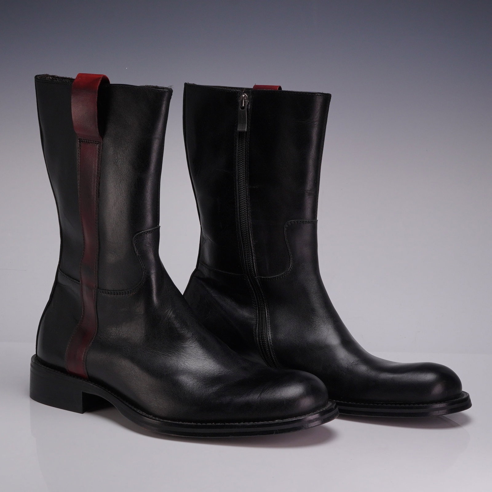 Pair of Kenneth Cole Vero Cuoio Black Leather Mid-Calf Side-Zip Boots: The boots are constructed with a sleek black leather upper characterized by a contrasting vertical burgundy leather strip on the lateral side that extends into a pull tab at the collar. The interior i