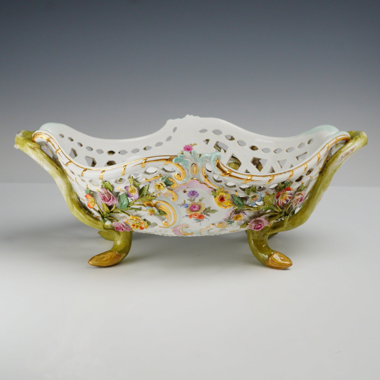 Meissen Reticulated Porcelain Floral Basket Centerpiece: A porcelain centerpiece basket featuring an elongated oval form with pierced reticulated sides, twin green branch-form handles, and four matching branch-form feet. The exterior is decorated with appli