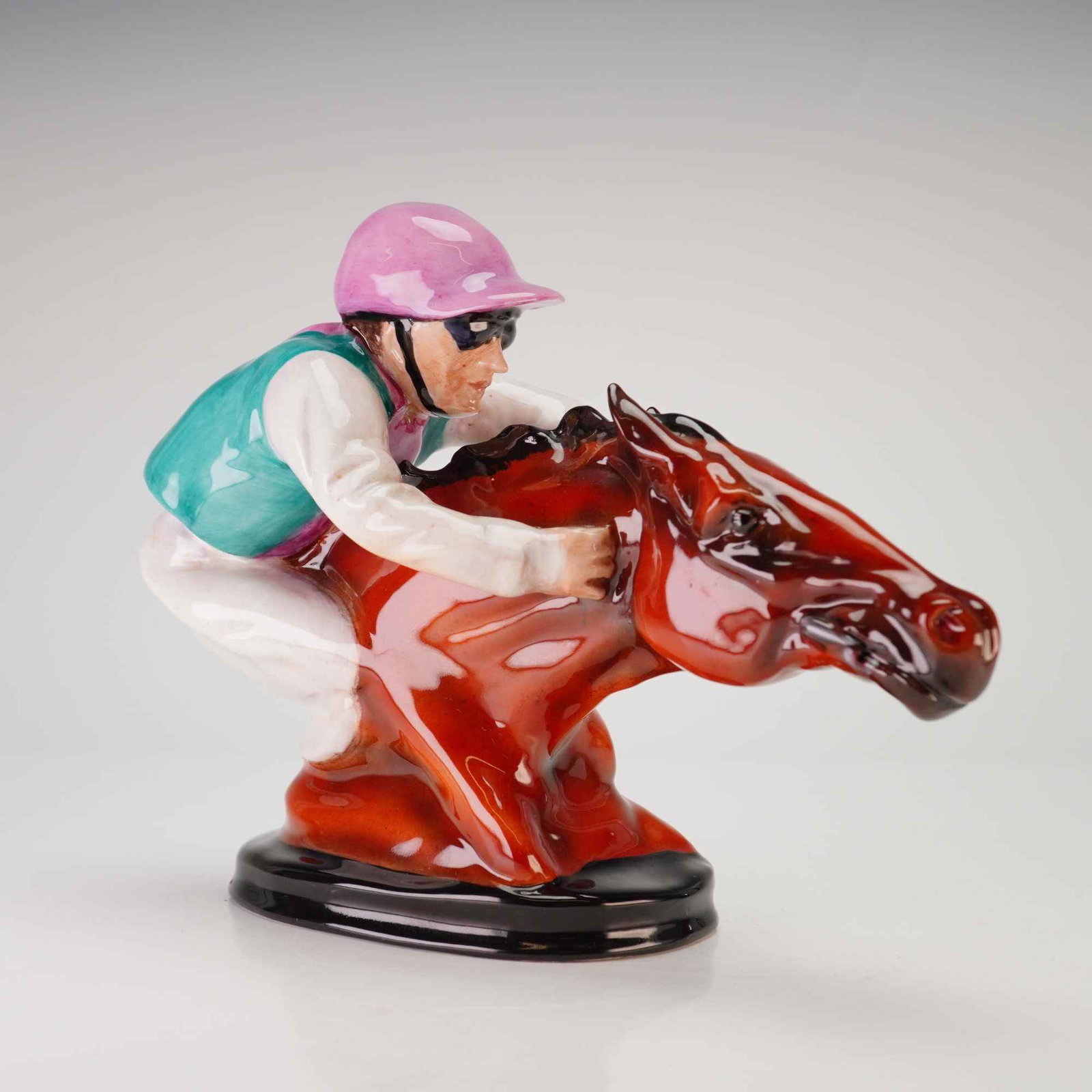 Michael Sutty Jockey and Horse Head Figurine: A Michael Sutty limited edition fine china figurine depicting a jockey in racing silks crouched atop the head and neck of a horse. The hand-painted porcelain piece features a jockey wearing a pink hel