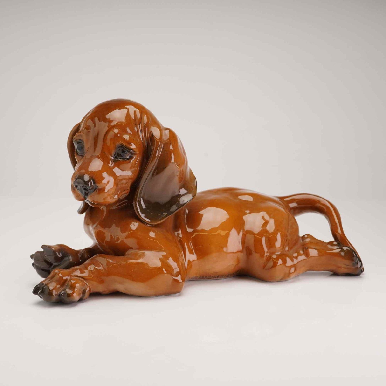 Rosenthal Porcelain Dachshund Puppy Figurine: A glazed porcelain figurine of a reclining dachshund puppy produced by the Rosenthal factory. The piece is rendered in a naturalistic style with a high-gloss brown glaze and hand-painted darker shaded