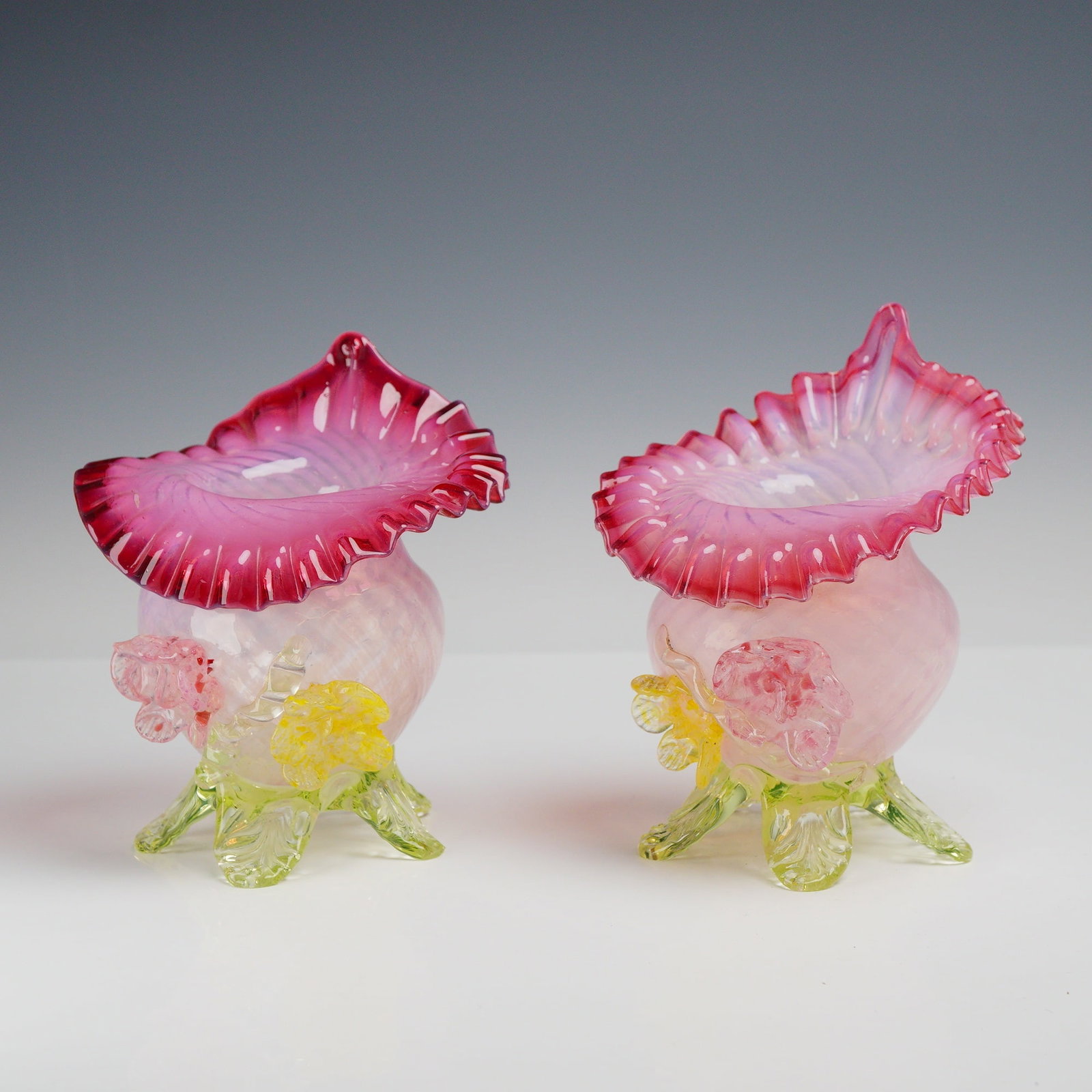 A Pair of Victorian Style Blown Art Glass Leaf Footed Uranium Ruffled Vases: A pair of hand-blown art glass vases in the Victorian style. Each vessel is constructed from cased, opalescent glass with a body that graduates from a pale pink to a deep ruby or cranberry red at the 