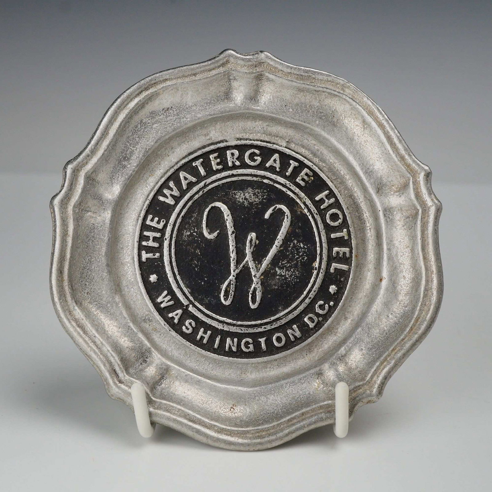 Vintage Historical Collectible Watergate Hotel Washington D.C. Pewter Ashtray: A vintage cast pewter ashtray or small souvenir dish from The Watergate Hotel in Washington, D.C. The piece features a scalloped molded rim with raised borders and a recessed black center medallion. T