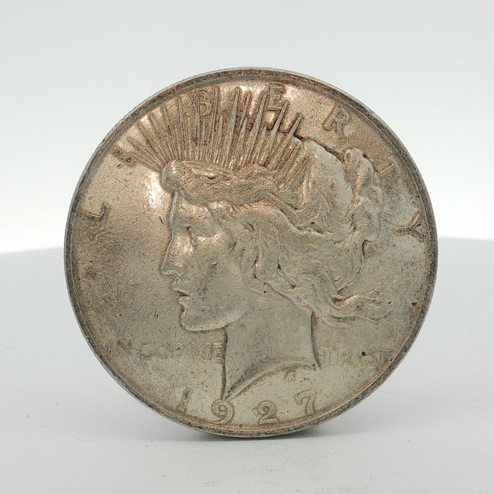 1927-D US One Dollar Silver Peace Dollar, Circulated: This coin was struck at the Denver Mint and is composed of .900 fine silver (90% silver, 10% copper). It features the standard Peace dollar obverse and reverse designs. The D mint mark is located on t