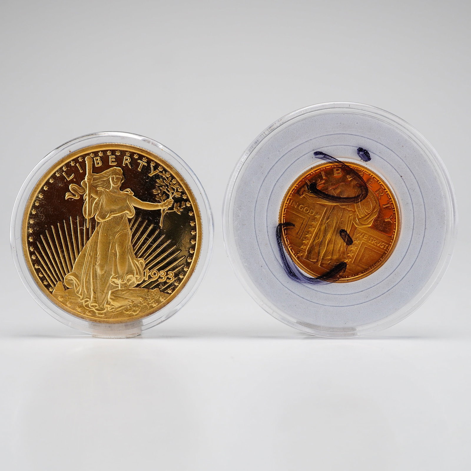 2pc Reproduction Coins, 1933 Saint-Gaudens Double Eagle Silver Bullion with Gold-Plating and Golden: This lot features two metal coins individually presented in clear plastic capsules. The first item is a silver bullion with gold-plating replica of a 1933 US Saint-Gaudens Double Eagle. The obverse fe