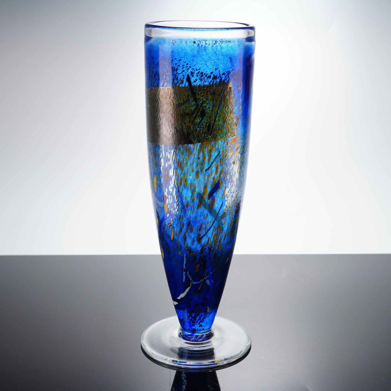 Kosta Boda by Bertil Vallien Art Glass Vase, Satellite: The piece is constructed of translucent cobalt blue glass in a tapering cylindrical form, terminating in a wide, clear circular foot. The decoration includes internal multi-colored frit in yellow, ora