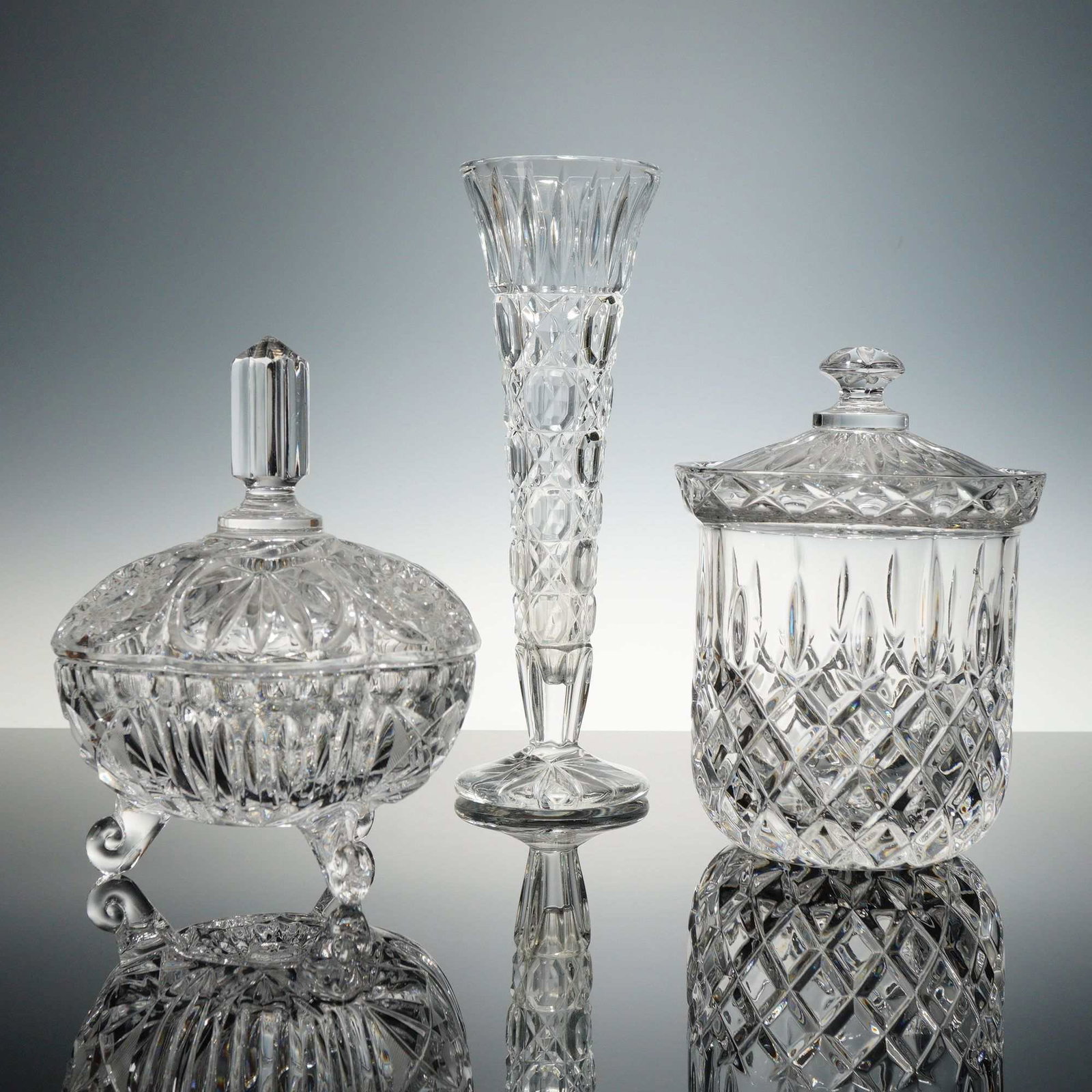 A Trio of Cut Crystal Decorative Tableware Including a Vase, Lidded Bowl, and Jar: This collection consists of three pieces of cut lead crystal featuring deep mitre cuts and geometric patterns. The first item is a footed lidded candy dish with a round bowl supported by three scrolle