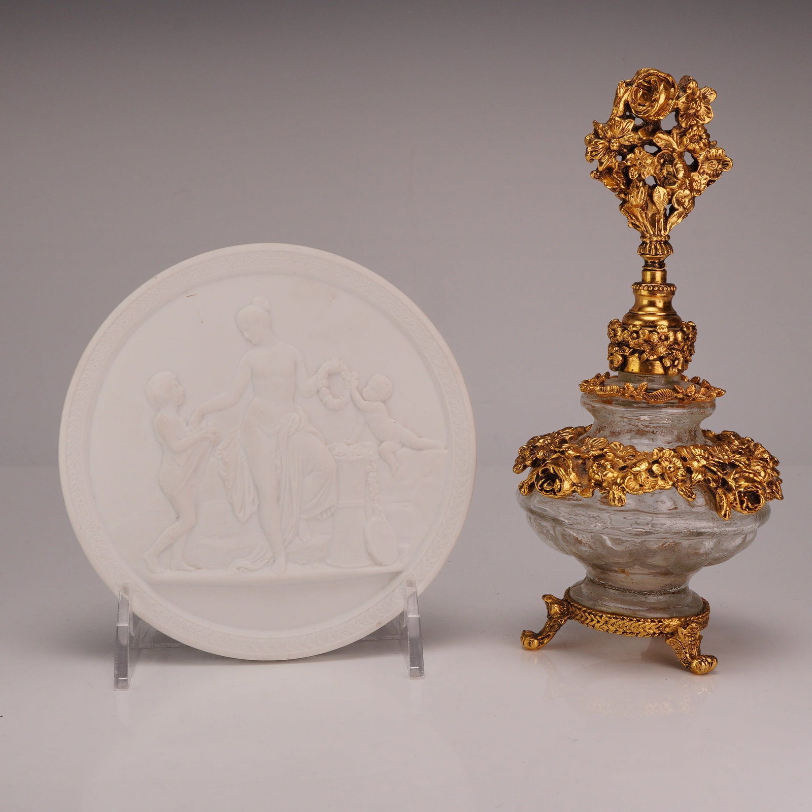 2pc Royal Copenhagen Bisque Porcelain Plaque & Floral Ormulu Mounts Perfume Bottle: This lot features a Royal Copenhagen bisque porcelain plaque and a vintage decorative ornate glass perfume bottle with a gold finish. The circular plaque was made in Denmark and features a bas-relief 