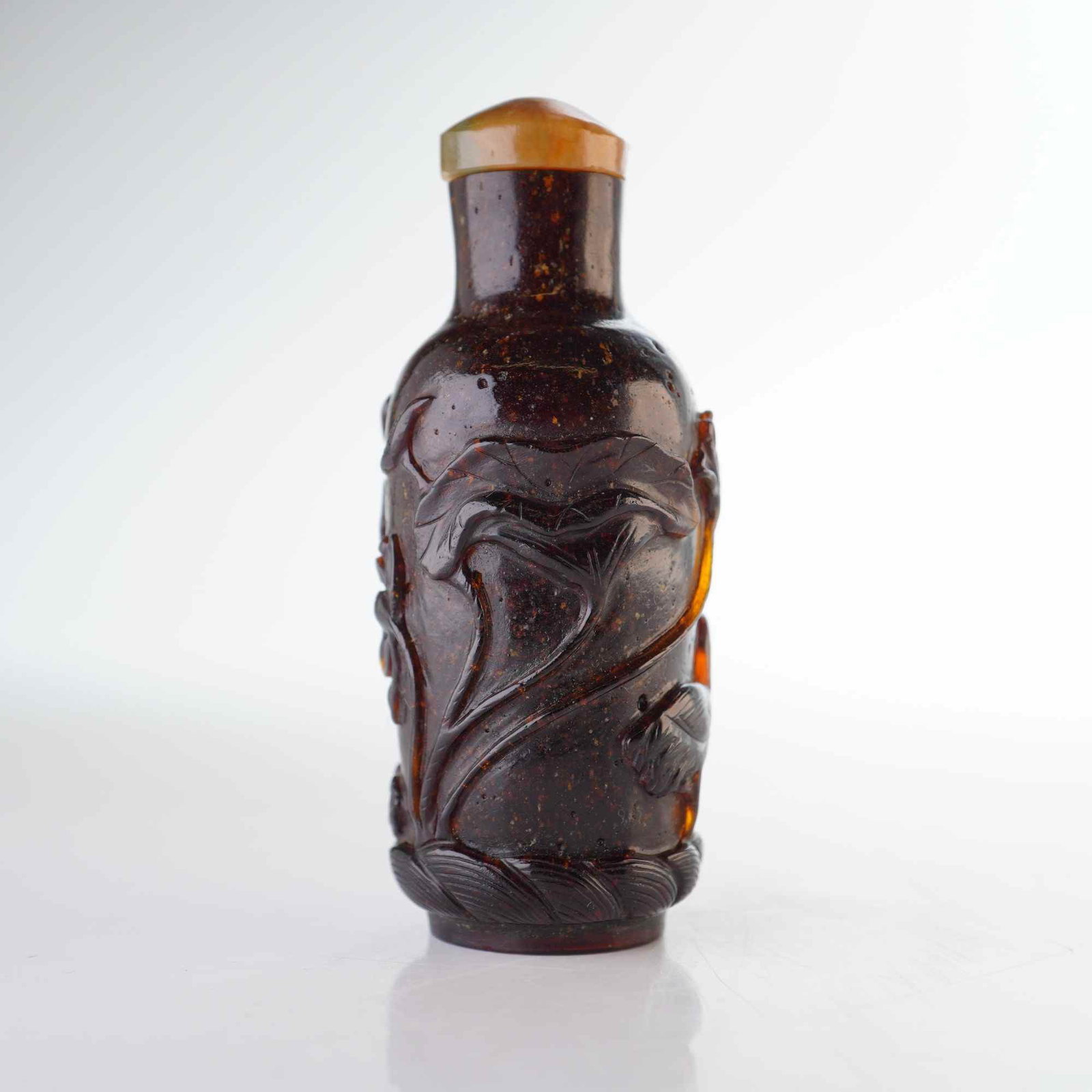 Peking Glass Snuff Bottle, Dark Amber Color, Lotus, Crane, and Ducks: A cylindrical Peking glass snuff bottle in a dark amber hue with a speckled pattern, featuring reliefs of lotus flowers, lotus leaves, cranes, and ducks. It is presented with a mottled tan, stone-like