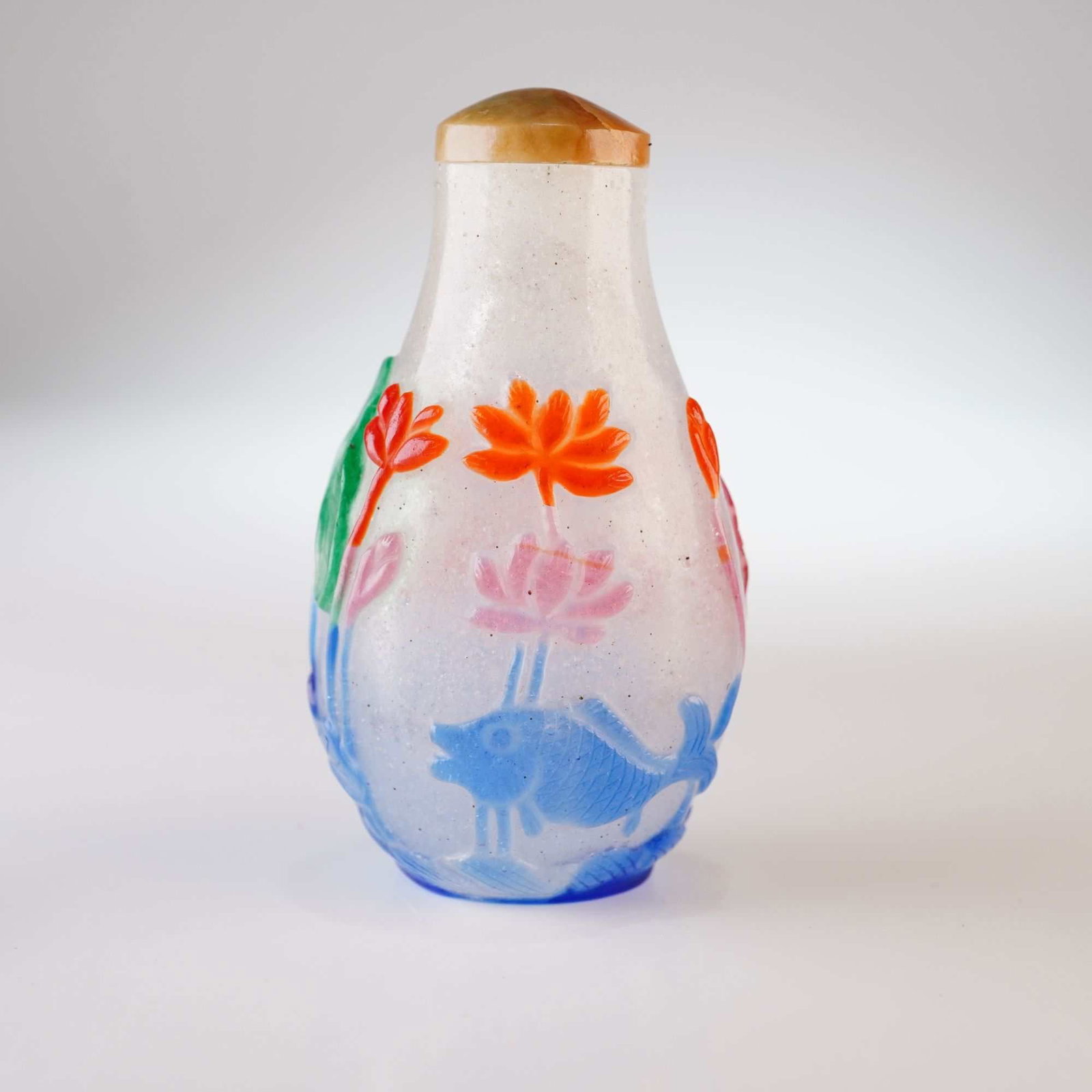 Peking Glass Snuff Bottle, Multi-color Overlay Fish and Lotus: This clear Peking glass bottle features a snowflake pattern and is decorated with red, blue, green, and pink relief overlay depicting lotus flowers, lotus leaves, and a fish. It is topped with a mottl