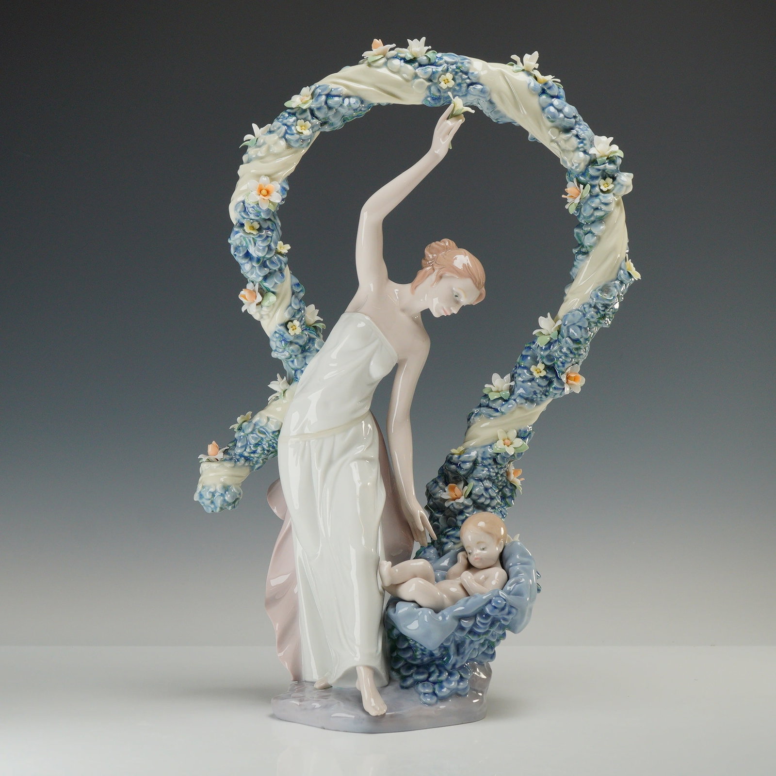 Lladro Inspiration Millennium Porcelain Figurine Rebirth 01006571 with Original Box: The sculpture depicts a woman in a white strapless gown standing beneath a circular garland arch composed of blue and white blossoms. She reaches upward toward the center of the arch. An infant is pos