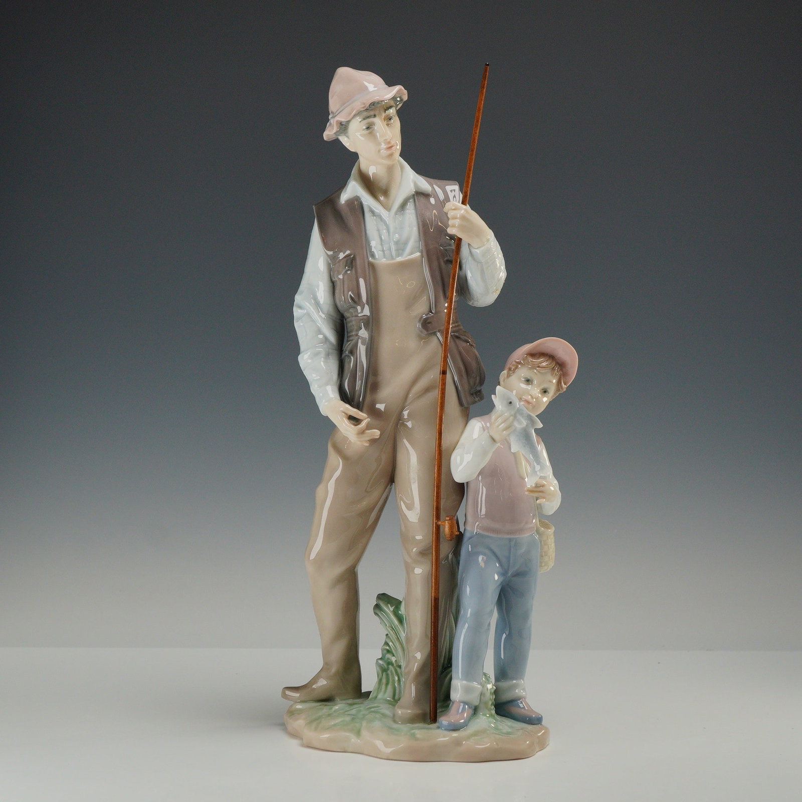 Lladro Porcelain Figurine A Father's Pride 01006467: The composition depicts an adult male and a young boy on a fishing outing. The adult figure is modeled wearing tan chest waders, a brown multi-pocket fishing vest over a light-colored collared shirt, 