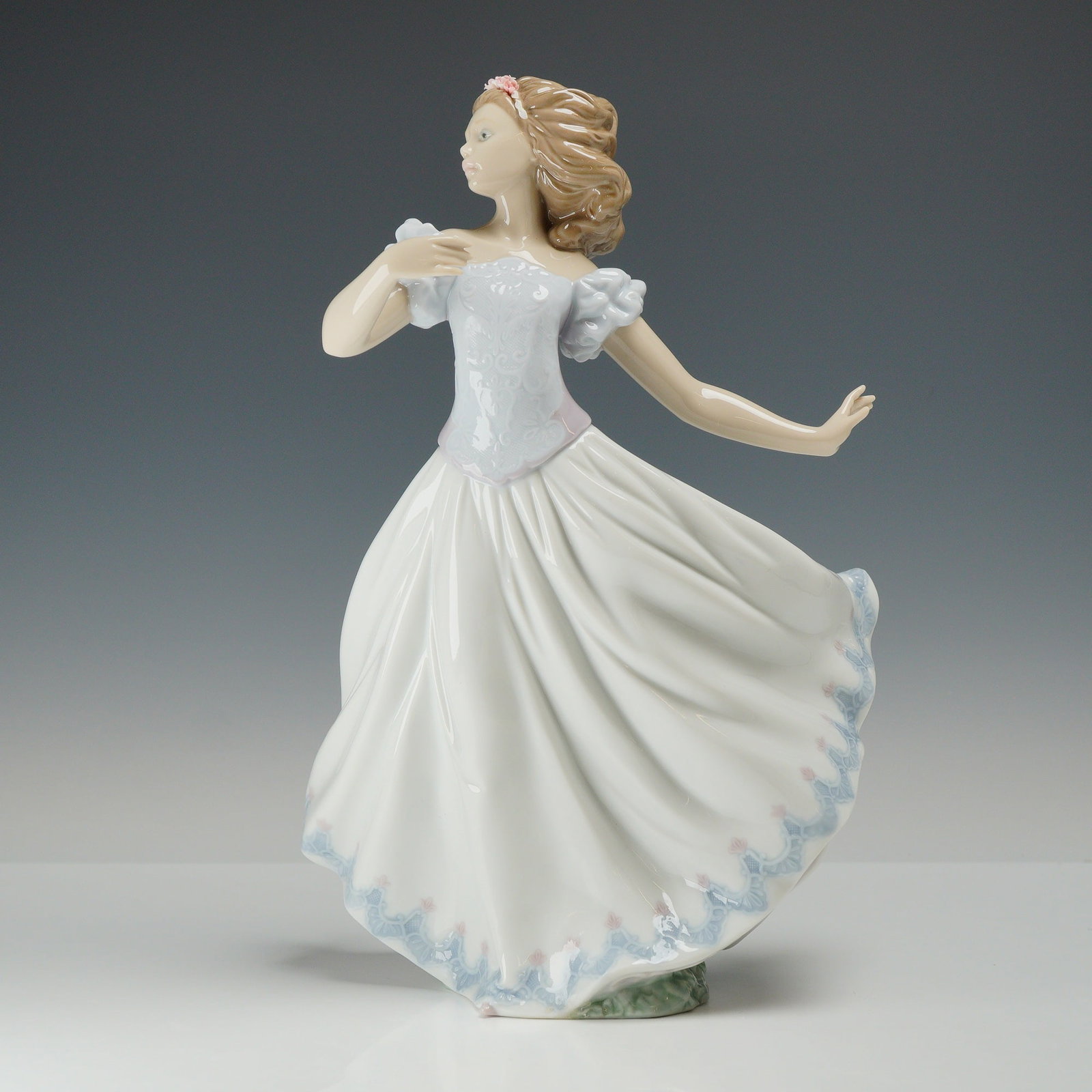 Lladro Porcelain Figurine Sweet Sixteen 01006687 with Original Box: The piece depicts a young woman in a dynamic pose, wearing a long, flowing white ballgown. The gown features a lavender-pink bodice and pale blue lace-style detailing along the scalloped hem. Her hair