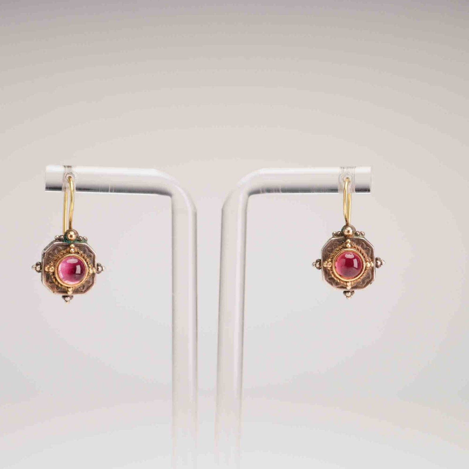 Pair of Sterling Silver Garnet Cabochon Drop Earrings: A matched pair of mixed-metal drop earrings featuring a Greek artisan design. Each earring is centered with a round cabochon-cut garnet, bezel-set within a yellow metal frame. The setting is accented 
