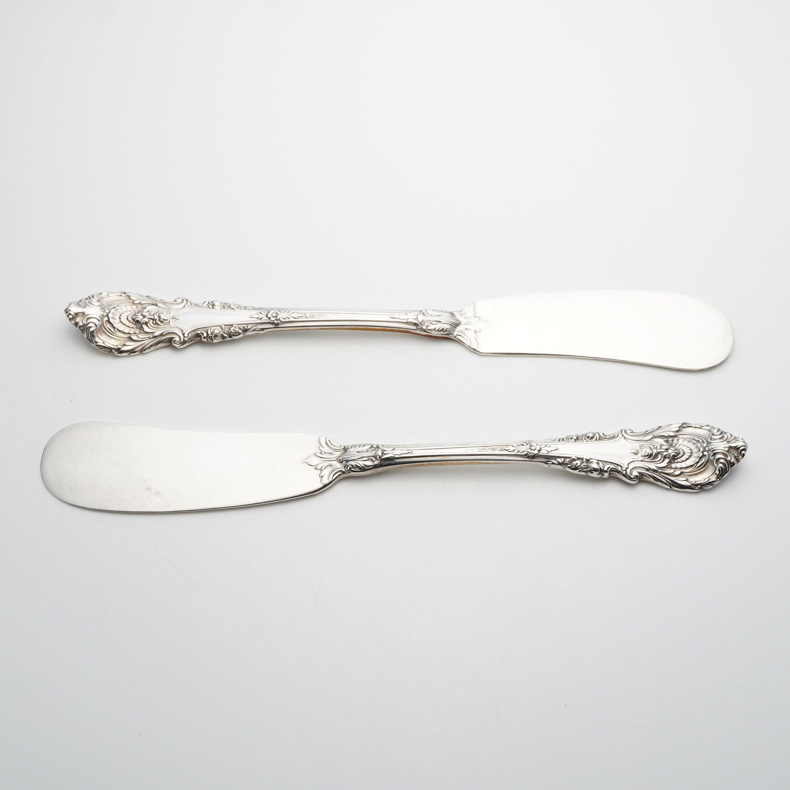 A Pair of Wallace Sir Christopher Sterling Silver Flat Handle Butter Spreaders: A pair of sterling silver butter spreaders in the Sir Christopher pattern by Wallace Silversmiths. These pieces feature a flat handle construction with a wide paddle-shaped blade. The handles are deco