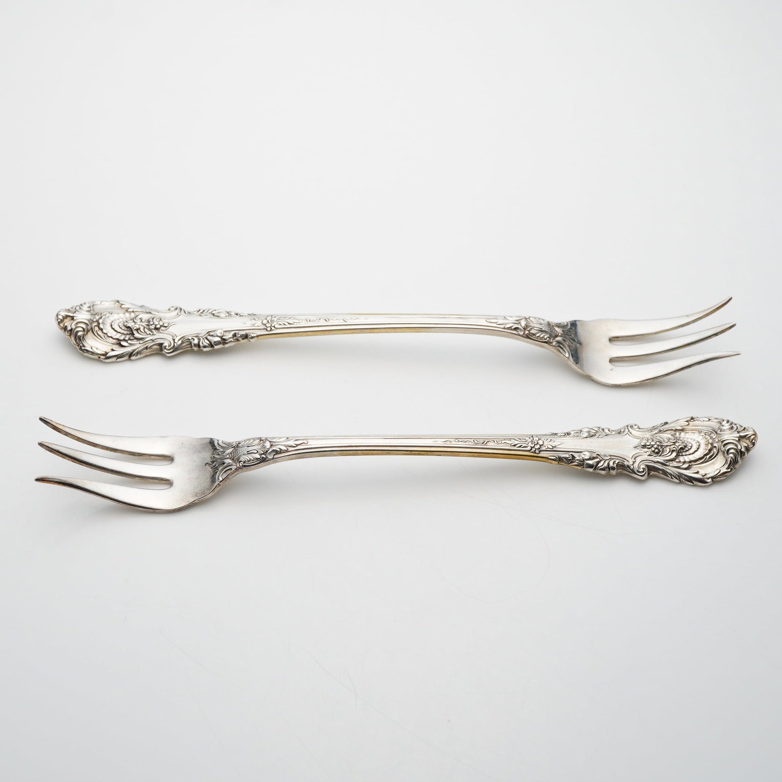 A Pair of Wallace Sterling Silver Cocktail Forks in the Sir Christopher Pattern: A pair of sterling silver cocktail or seafood forks manufactured by Wallace Silversmiths in the Sir Christopher pattern. Introduced in 1936, this pattern features an elaborate design inspired by the E