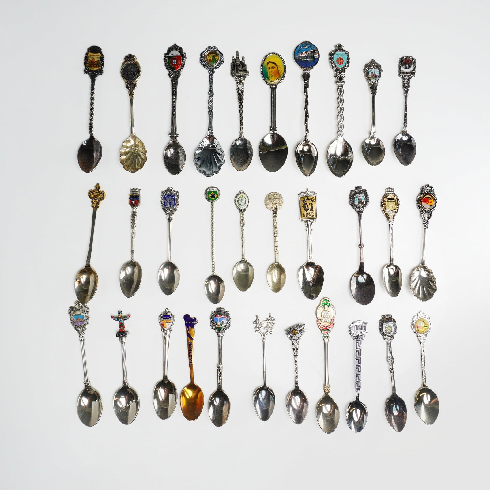 31pc Commemorative Souvenir Sugar and Tea Spoons: This collection consists of 31 commemorative souvenir sugar and tea spoons crafted from various metals, including silver-plate and one gold-toned example. The spoons feature a diverse range of decorat