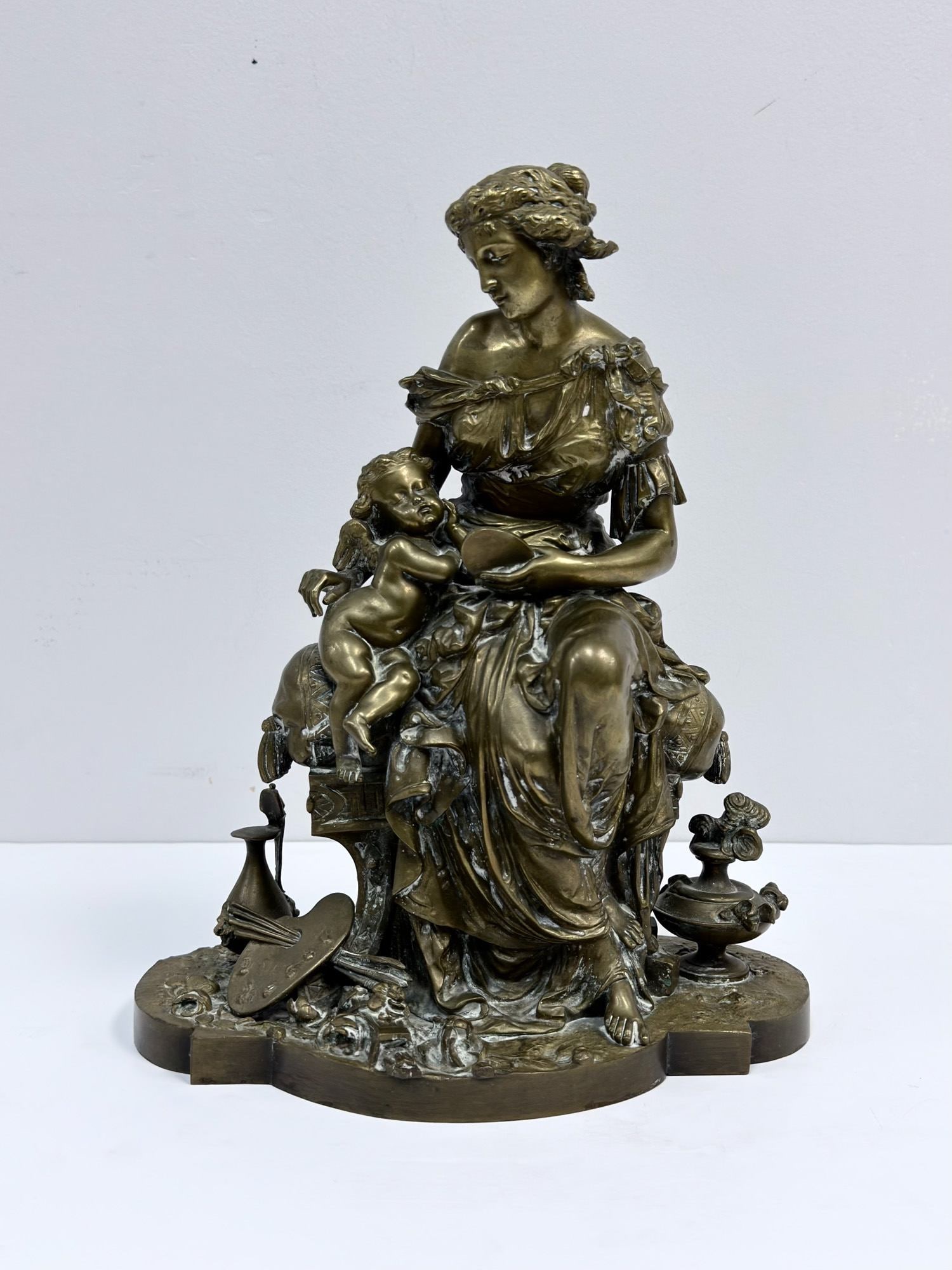 Dhumege Patinated Bronze Sculpture of a Classical Maiden and Cupid: Depicting an allegorical scene of a seated classical maiden and a winged putto, or Cupid. The woman is dressed in flowing, Greco-Roman style drapery and is seated upon a tasseled, cushioned bench feat