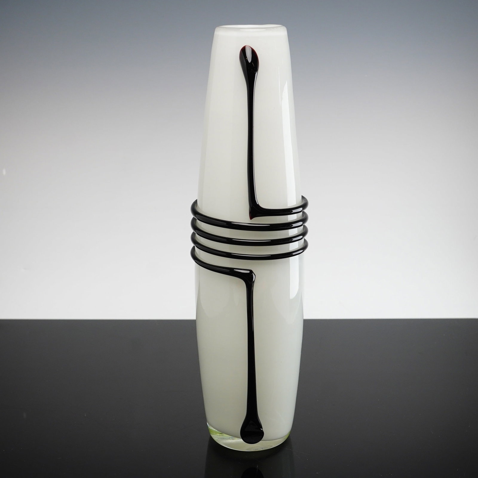 Attributed to Jerzy Sluczan-Orkusz, Art Glass Vase with Black Coil Decoration: This mid-century modern art glass vase is attributed to Polish designer Jerzy Sluczan-Orkusz. The vessel features a tapering cylindrical form crafted from an opaque white milky glass body cased in a l