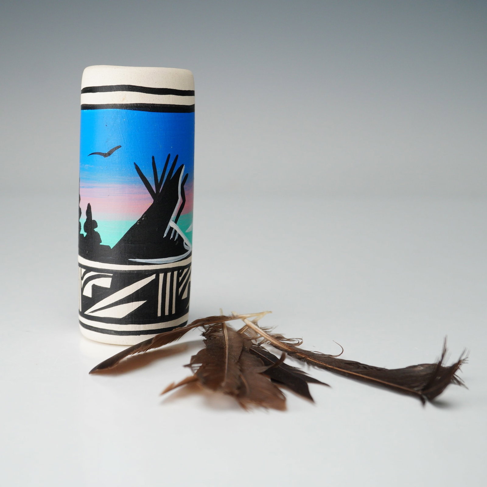 Native American Hand-Painted Feathered Pottery Vase: Hand-painted cylindrical pottery vase decorated with vivid gradient bands of blue, pink, and green, accented with bold black silhouetted scenes of tipis, riders on horseback, and soaring birds. The ba