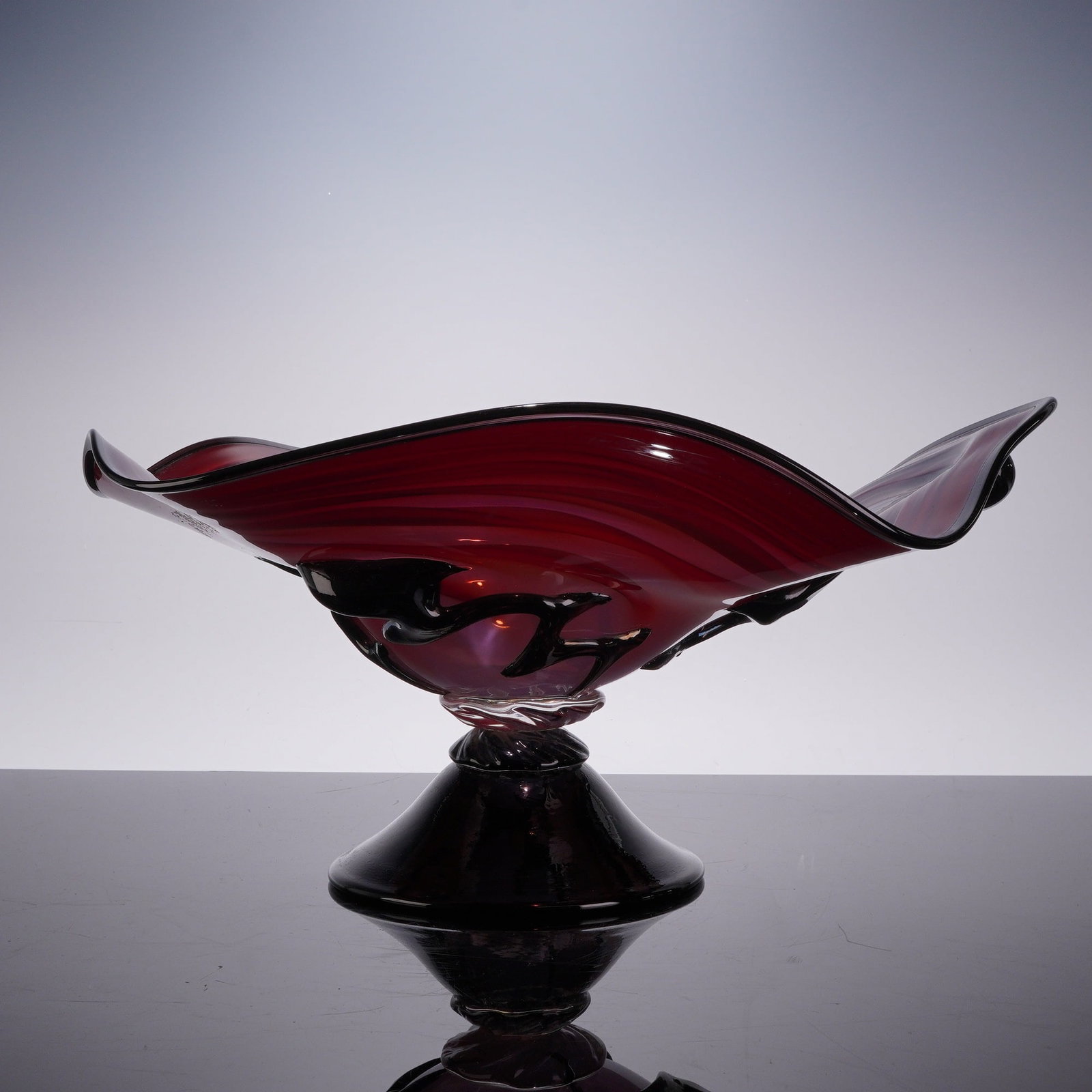 C. Hall Hand-Blown Art Glass Centerpiece Bowl: The contemporary vessel is executed in reddish purple glass featuring a wavy pattern of horizontal swirled striations in shades of dark purple and black. The bowl is designed with an asymmetric, undul