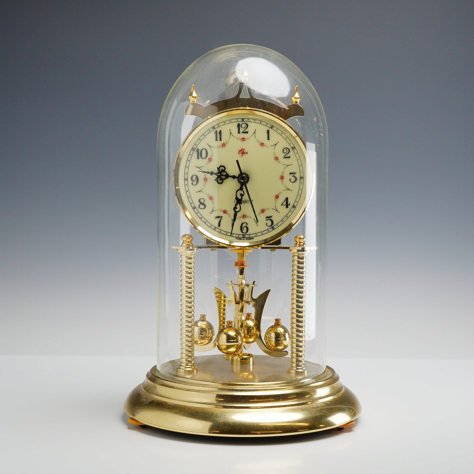 Elgin Quartz Anniversary-Style Mantel Clock with Glass Dome: This is a quartz anniversary-style mantel clock manufactured by Elgin. The clock features a polished brass-toned metal base with two fluted vertical pillars supporting the central movement. The cream-
