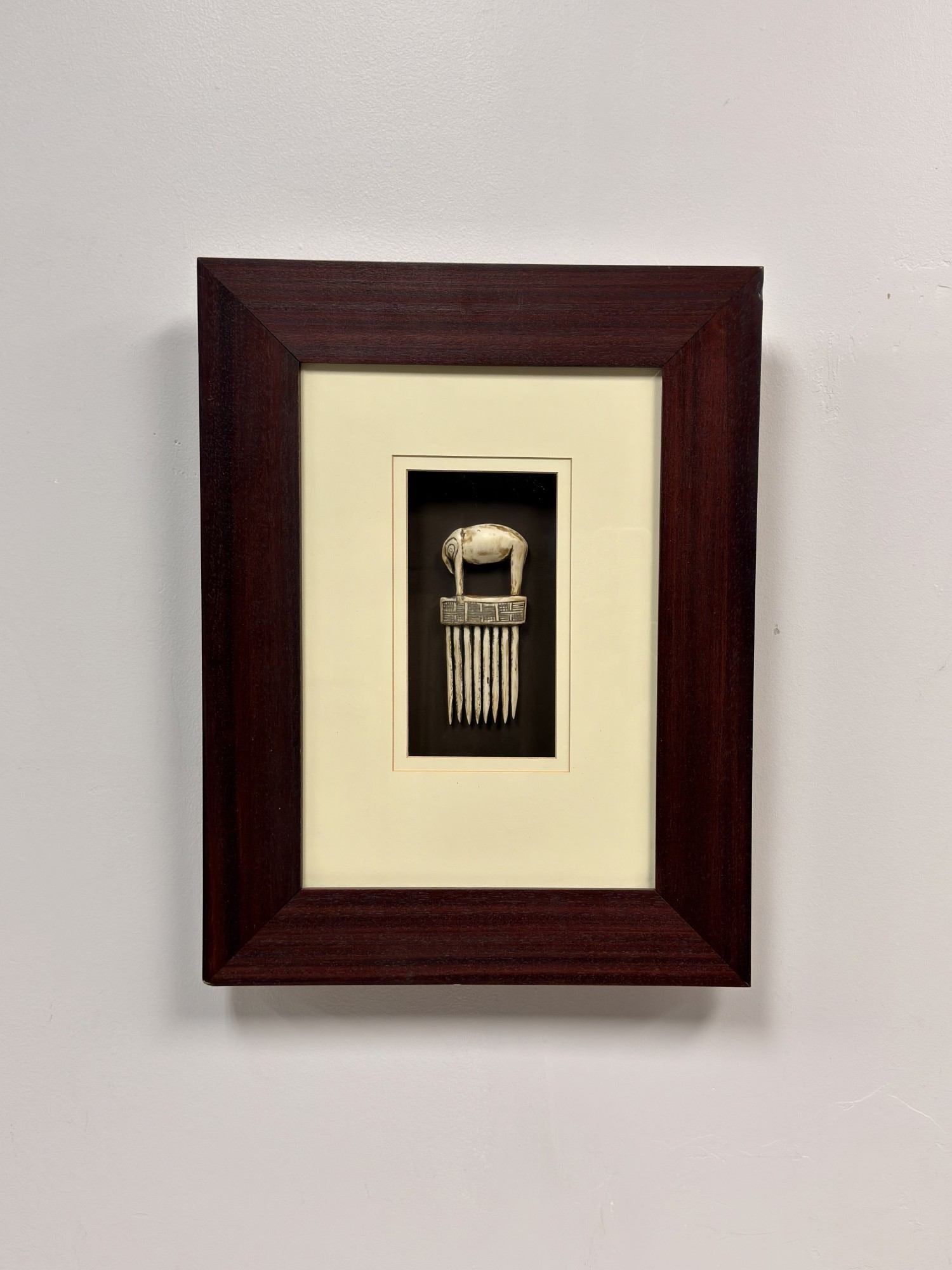Framed Carved Bone Comb with Elephant Finial: This framed item is a decorative comb hand-carved from bone with a light-colored patina within the incised details. The handle is carved in the form of a standing elephant with its head and trunk lowe