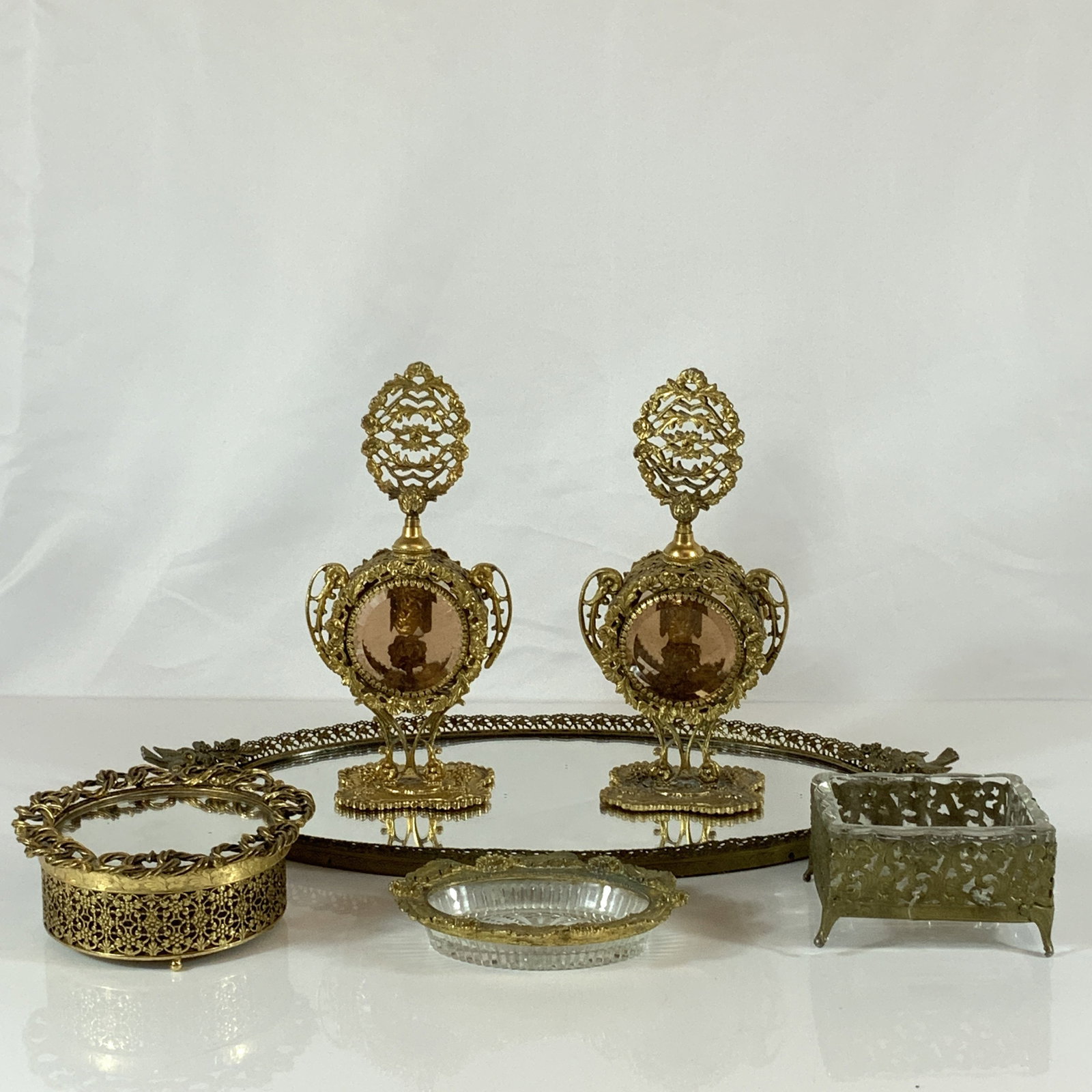 6pc Glass and Gold Tone Vanity Collection in the Hollywood Regency Style: This 6pc glass and gold tone vanity collection is designed in the Hollywood Regency style, featuring ornate gold tone metalwork and decorative glass elements. The collection includes a large oval mirr