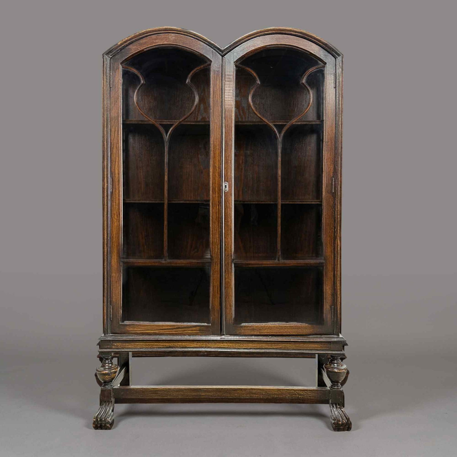 Early 20th Century Double Arch Oak Display Cabinet: This is a double arch oak display cabinet on stand dating to the early 20th century, designed in the William and Mary Revival style. The upper glazed section features a distinctive double-arched corni