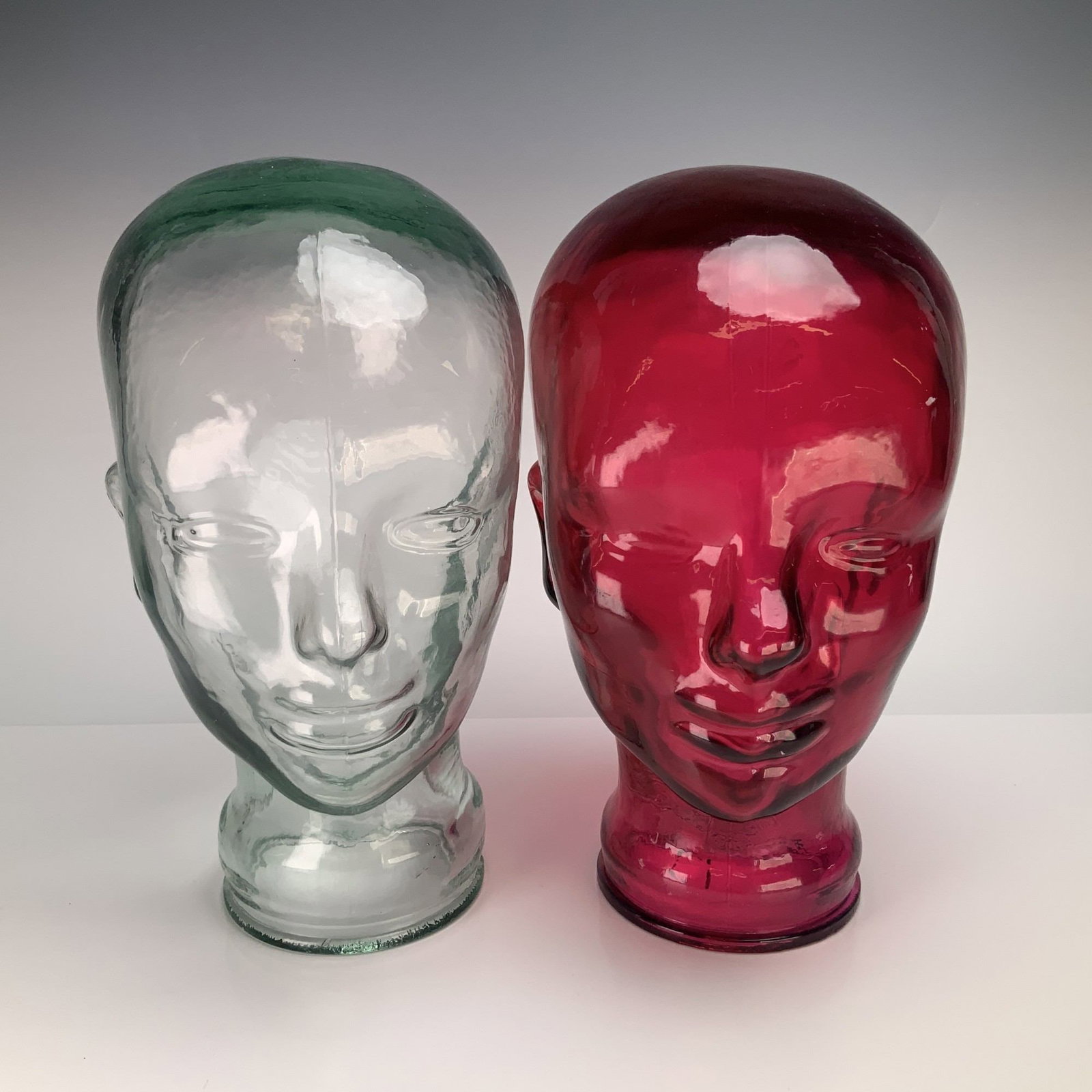 2pc Decorative Mold-Blown Glass Mannequin Heads in Deep Pink & Clear: A pair of life-size decorative mannequin heads crafted from mold-blown glass. The set includes one head in clear glass with a faint green tint and one head in a deep pink glass. Each head features sty