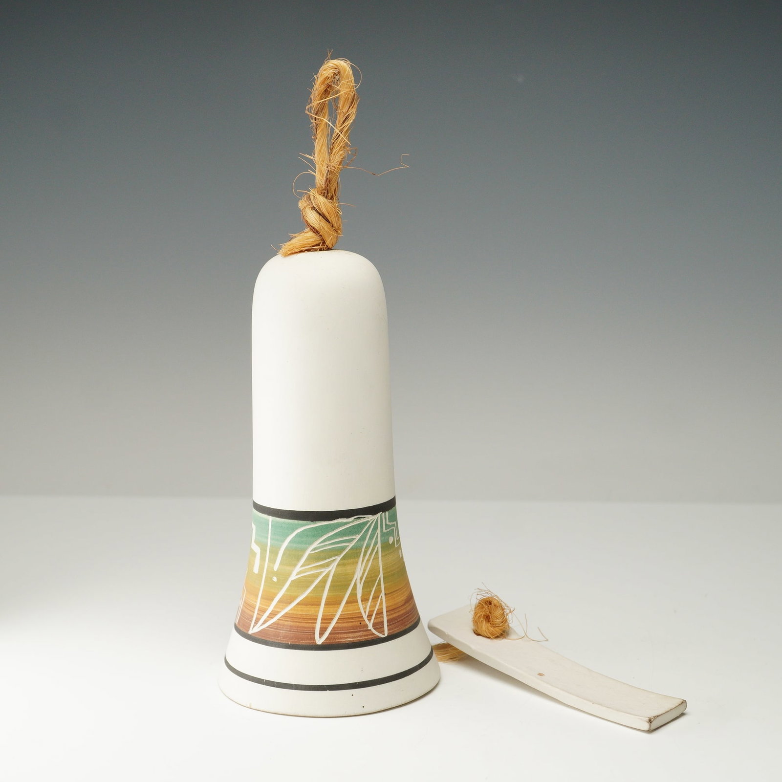 Navajo Incised Pottery Bell: Handcrafted Navajo pottery bell with a matte white surface, decorated with a band of incised geometric and leaf motifs over a multicolor gradient ground in warm earth tones. The bell features a rope h