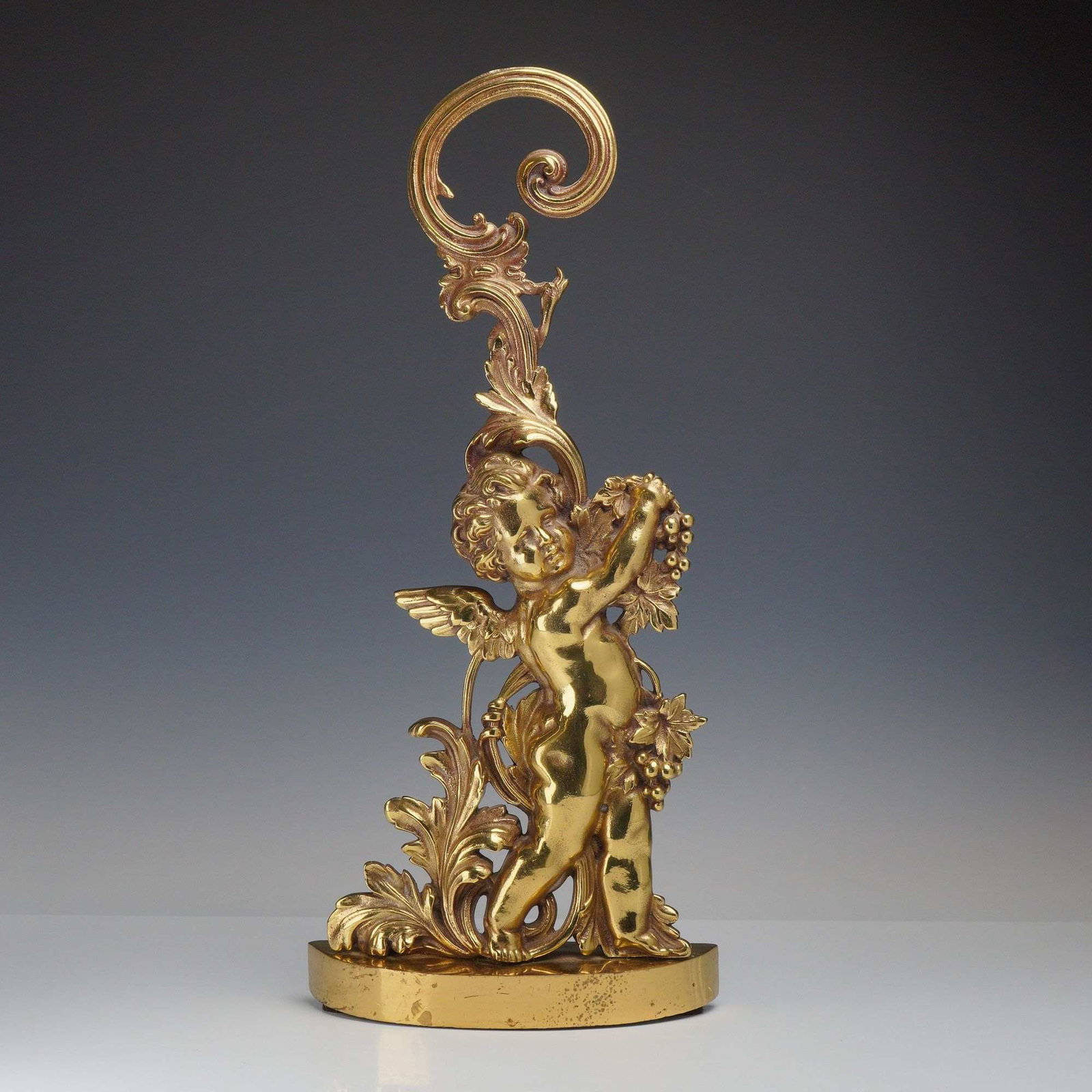 Peerage Cast Brass Cherub and Acanthus Scroll Doorstop: A vintage cast brass doorstop manufactured by Peerage of England. The piece is designed in the Rococo Revival style, featuring a central winged cherub or putto standing amidst elaborate scrolling acan