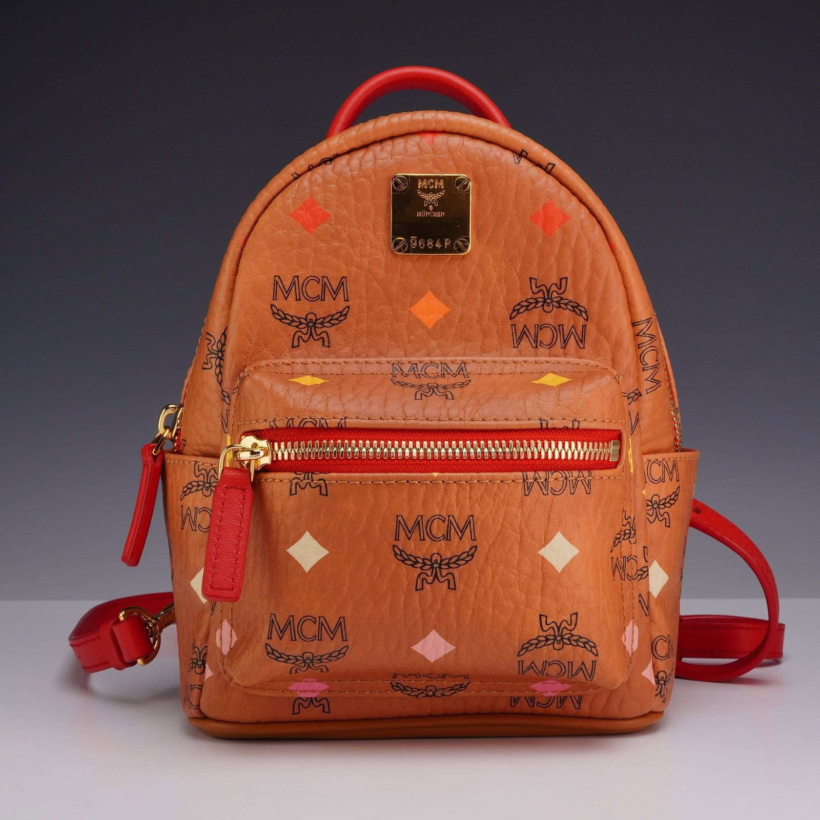 MCM Skyoptic Visetos X-Mini Stark Bebe Boo Backpack in Cognac and Red: This is an MCM Skyoptic Visetos X-Mini Stark Bebe Boo backpack, a convertible knapsack design. The exterior is crafted from signature cognac-colored Visetos coated canvas, featuring a unique multi-col