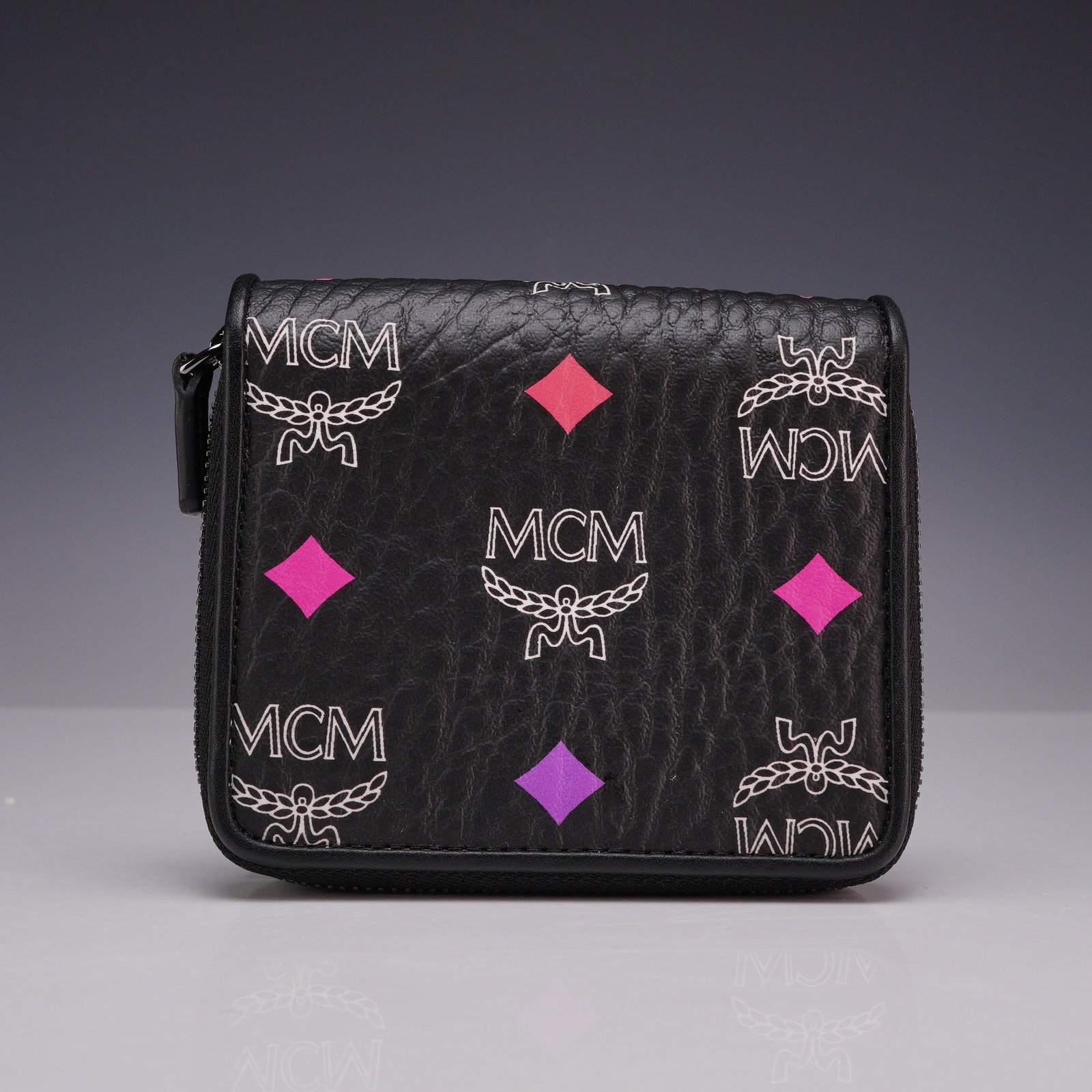 MCM Spectrum Diamond Visetos Bifold Zip-Around Wallet in Black: An MCM Spectrum Diamond Visetos bifold zip-around wallet crafted from black pebbled leather. The exterior features the signature Visetos monogram print in a white-toned outline, accented by diamond mo