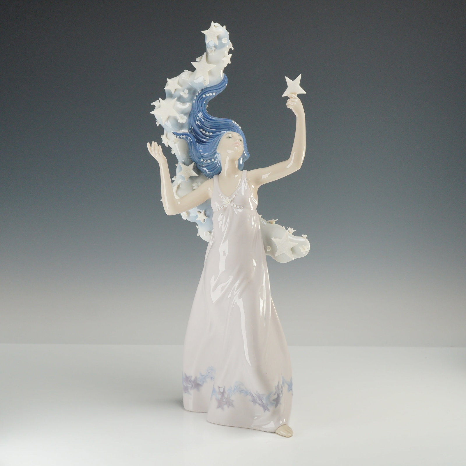 Lladro Inspiration Millennium Porcelain Figurine The Milky Way 01006569: The piece is crafted from glazed porcelain and depicts a stylized female figure in a long, flowing pale pink gown. The figure is shown with arms raised, holding a single five-pointed star in her left 