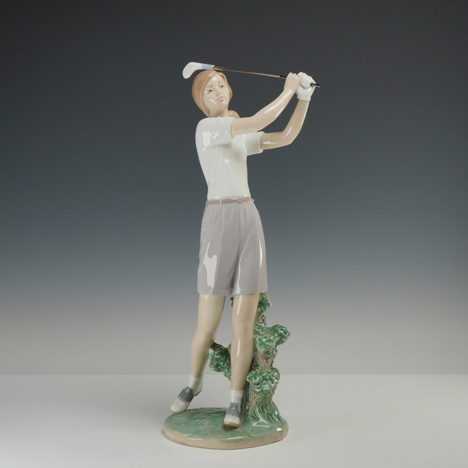 Lladro Porcelain Figurine A Perfect Drive 01006689: A Lladro glazed porcelain figurine titled A Perfect Drive depicting a female golfer in a follow-through swing. The figure is rendered in a palette of gray, white, and tan tones with a high-gloss finis