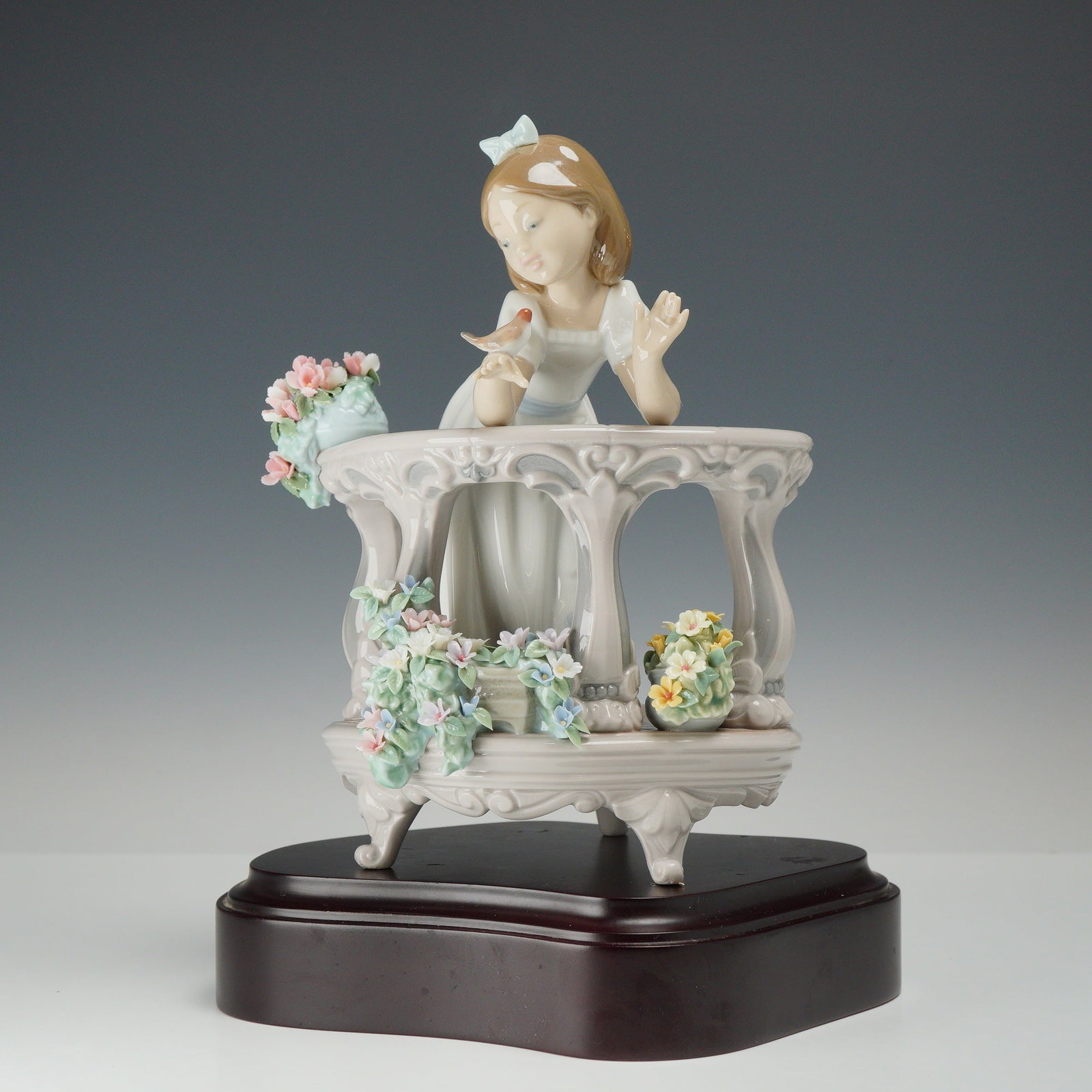 Lladro Porcelain Figurine Morning Song Girl 01006658: The piece depicts a young girl in a white gown with a light blue sash and matching hair bow, leaning over a classical-style balcony. The balcony is decorated with various encrusted floral arrangements