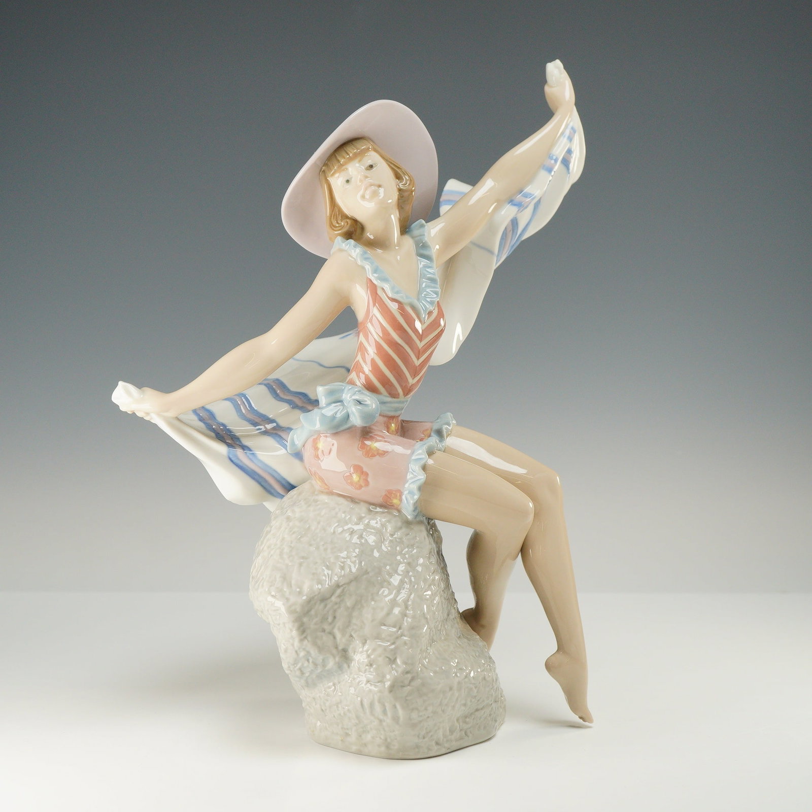 Lladro Porcelain Figurine Sunning in Ipanema 01005660: The figurine depicts a young woman in a stylized vintage-inspired swimsuit and a wide-brimmed lavender sun hat adorned with a bow, seated atop a textured, rock-like base. She is portrayed in a dynamic
