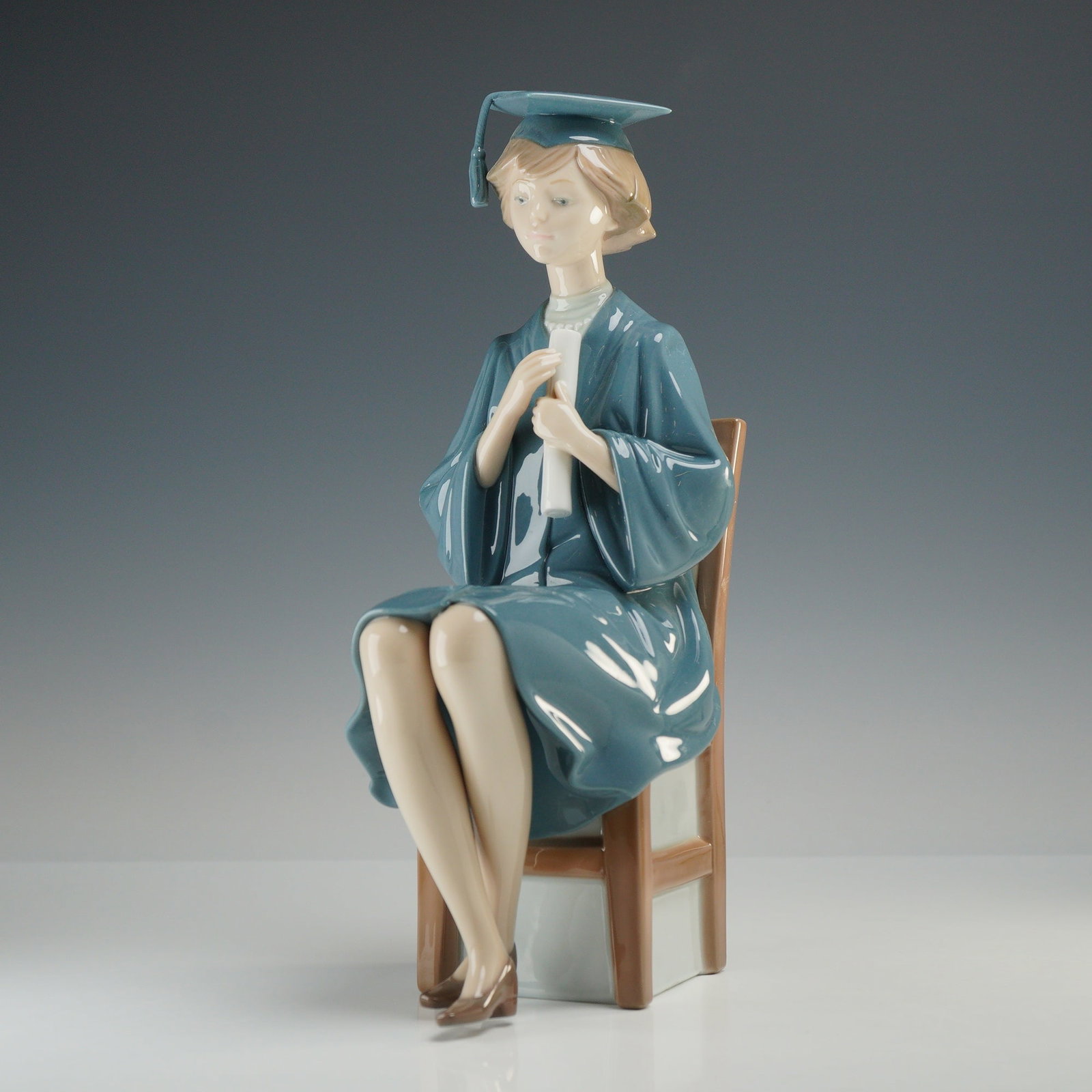 Lladro Porcelain Figurine, Girl Graduate 01005199: This Lladro porcelain figurine, titled "Girl Graduate" (model 01005199), depicts a seated young woman in graduation regalia holding a diploma. The figurine is exquisitely decorated in glazed porcelain