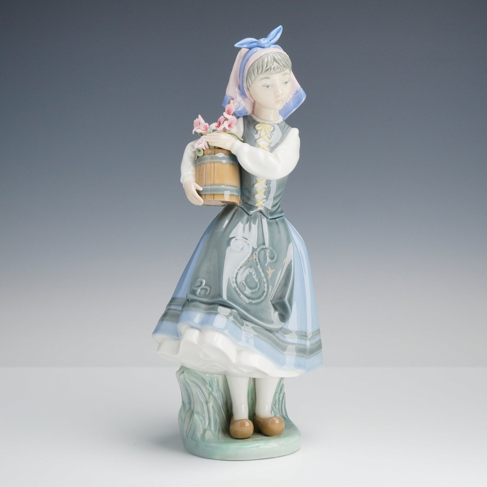 Lladro Porcelain Figurine, Budding Blossoms 1001416: A Lladro glazed porcelain figurine identified as Budding Blossoms, model number 1001416. The figure depicts a young girl in folk attire, featuring a light blue and white headscarf, a dark grey vest wi