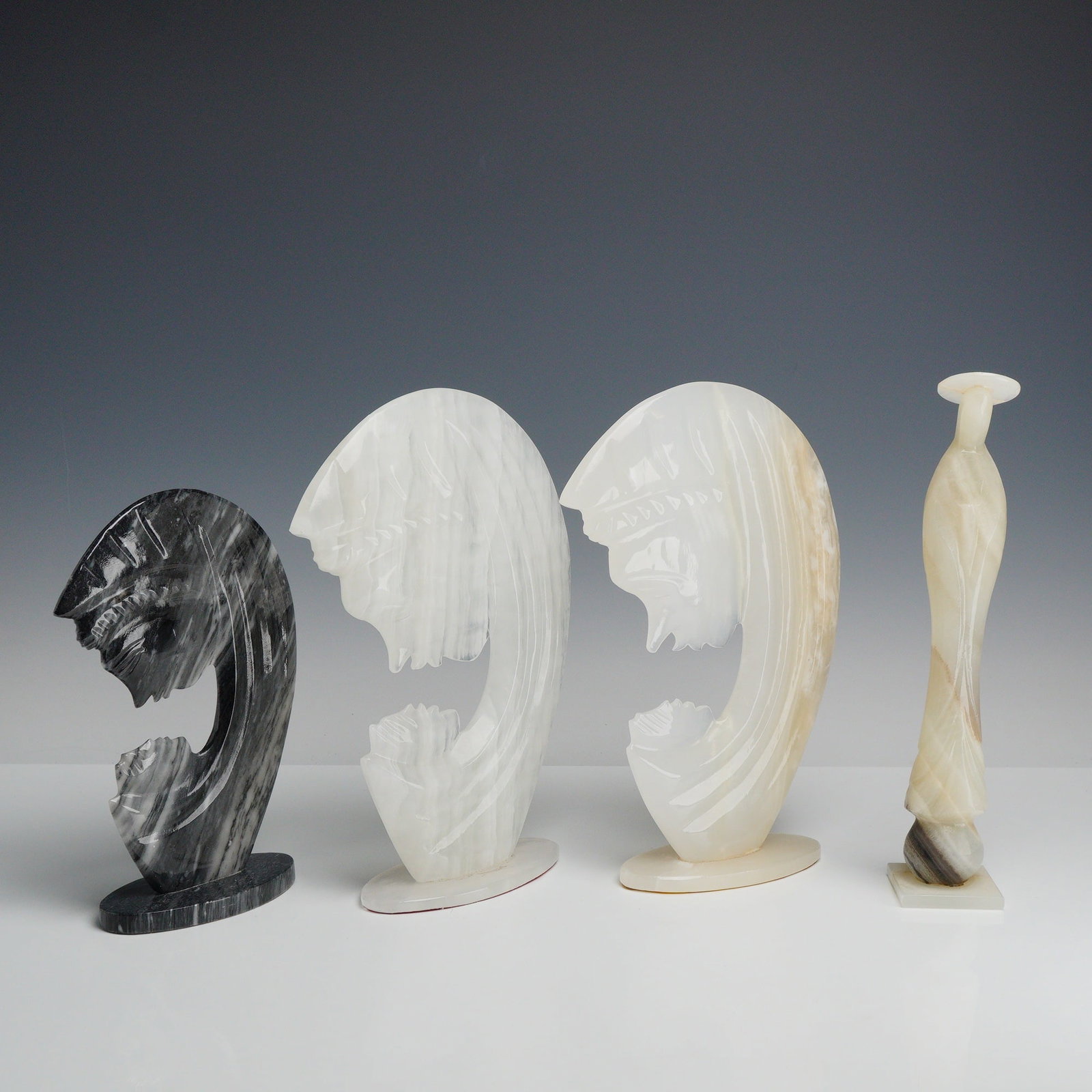 A Set of 4 Carved Stone Mother Mary Figures: A collection of four modernist carved stone sculptures representing Mother Mary figures. The group consists of three stylized profile busts and one attenuated female figure. The profile busts are unif