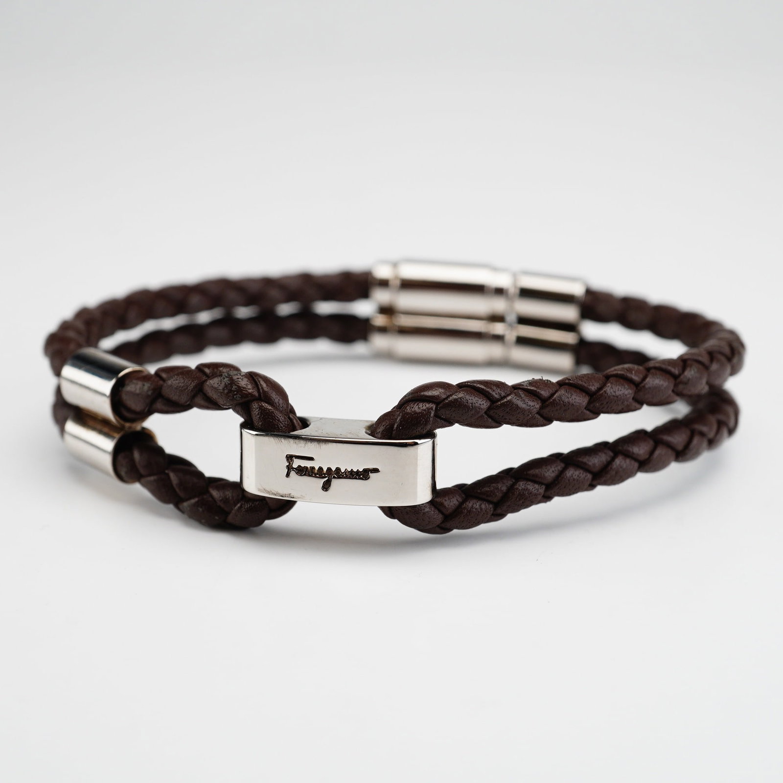 Ferragamo Italian Braided Leather Bracelet: Crafted in brown leather, this bracelet features a double-strand braided design, finished with polished silver-tone metal hardware and a magnetic closure. The hardware is engraved with the Ferragamo s