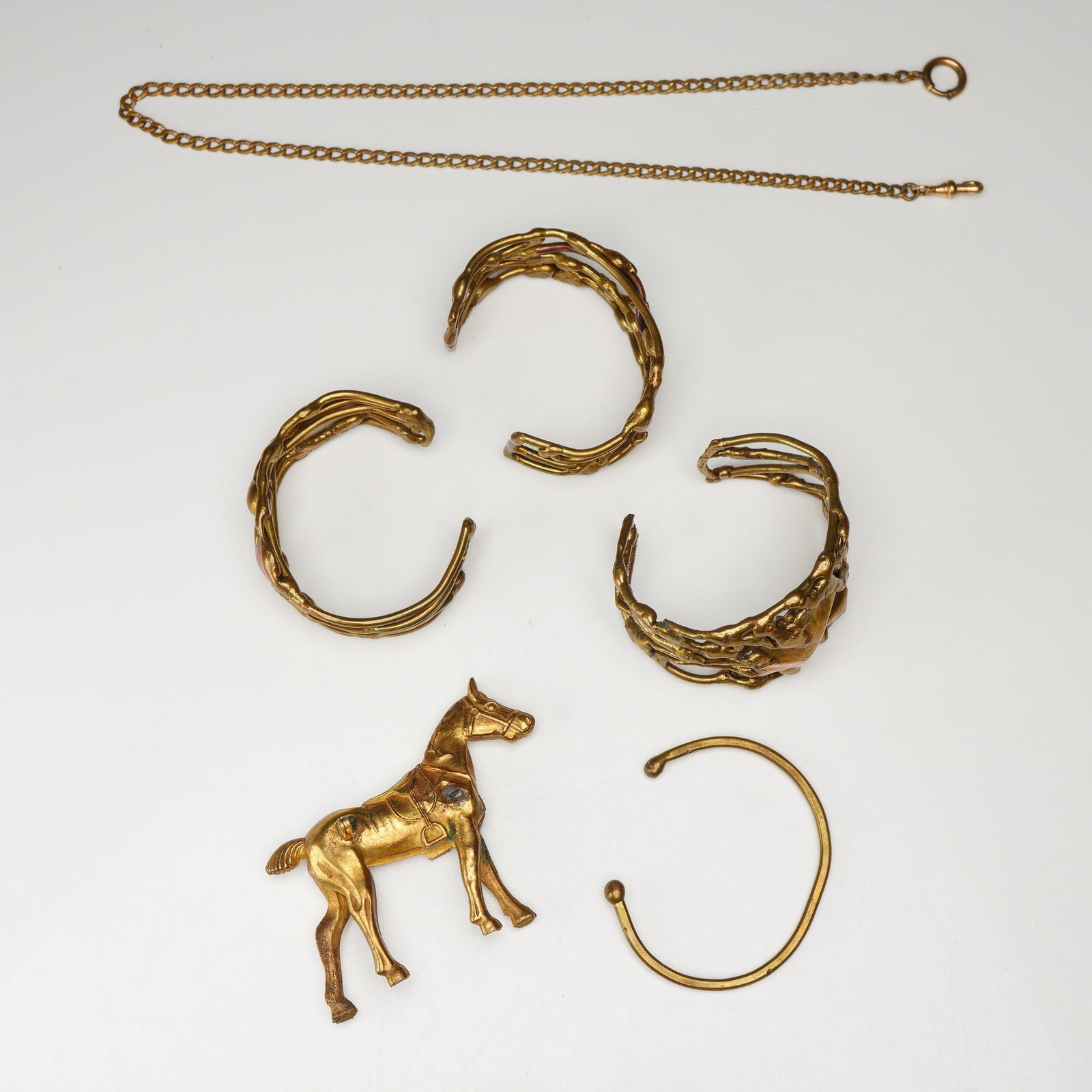 Six Brass Jewelry Including Four Cuff Bracelets, Horse Pin and a Watch Chain: Three of the cuff bracelets are designed in a Brutalist style, featuring an organic, molten-textured open-work design, while the fourth is a slender cuff with ball-shaped terminals. The gold-tone meta