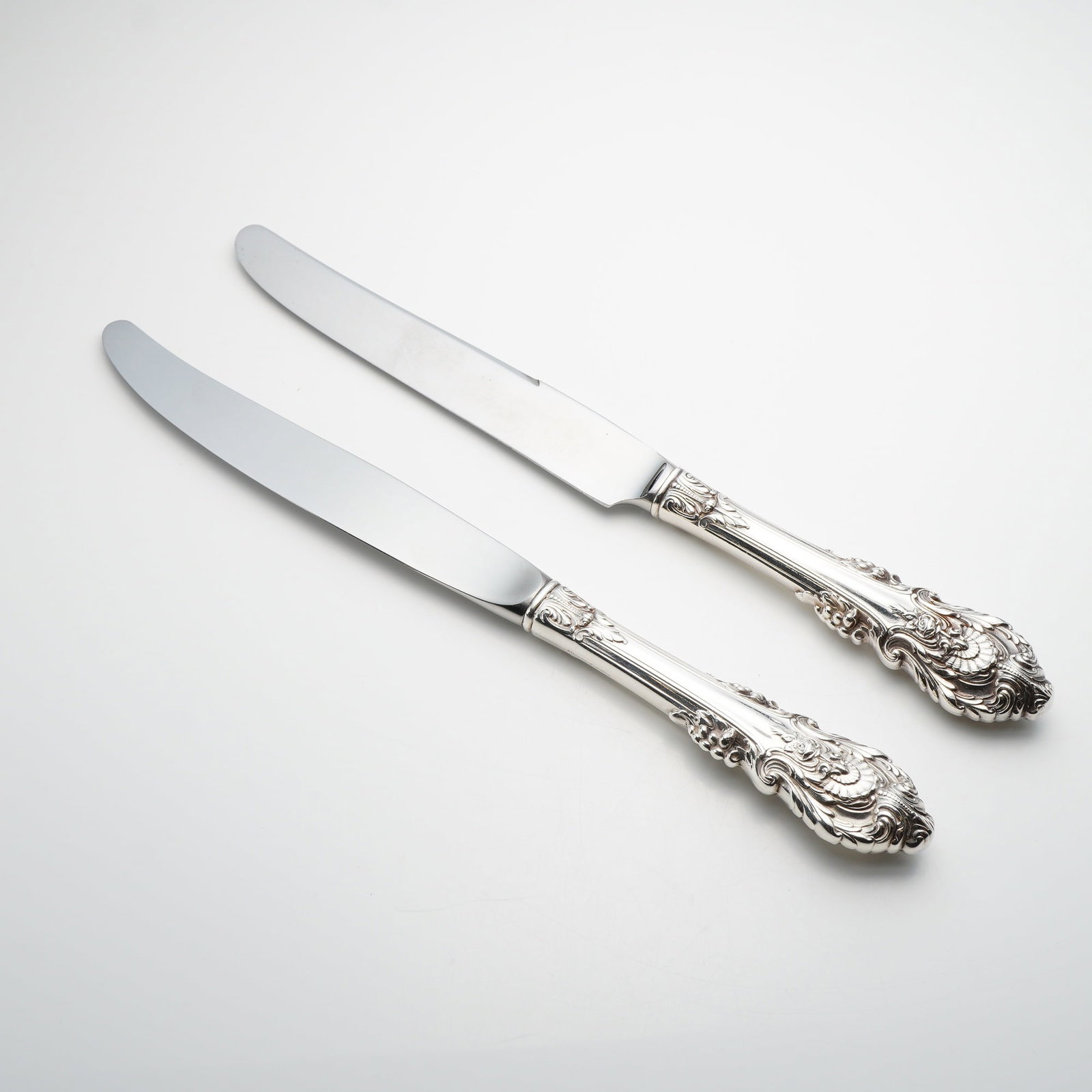 A Pair of Wallace Sterling Silver Sir Christopher Pattern Dinner Knives: This pair of dinner knives is manufactured by Wallace Silversmiths in the Sir Christopher pattern. Each piece features a weighted hollow handle crafted from sterling silver, decorated with intricate t