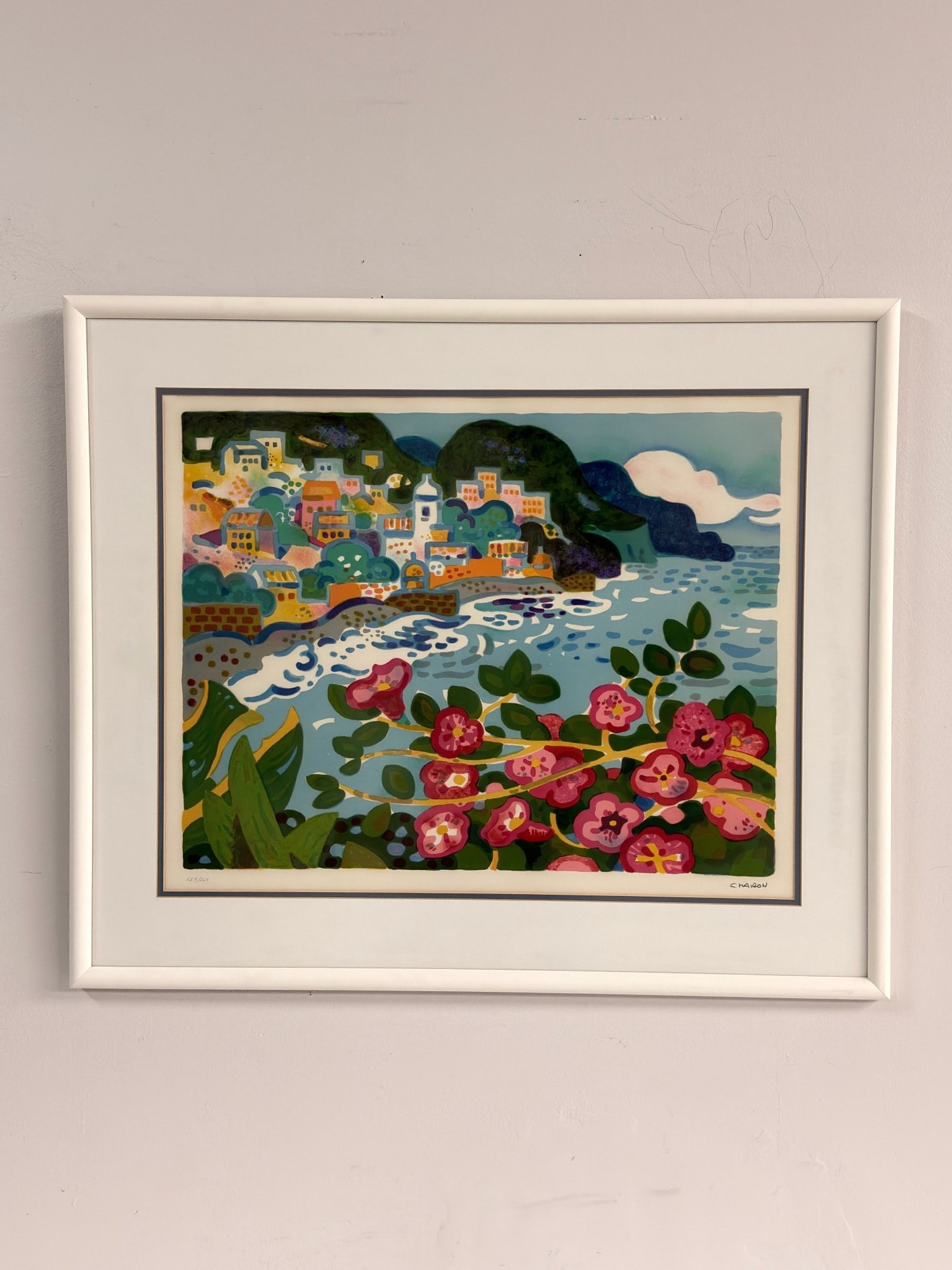 Guy Charon (French 1927-2021) Lithograph on Paper, Vibrant Coastal Scene, Signed: This lithograph by Guy Charon depicts a coastal landscape featuring a hillside village with multicolored architecture overlooking a shoreline. The foreground is composed of pink floral motifs and gree
