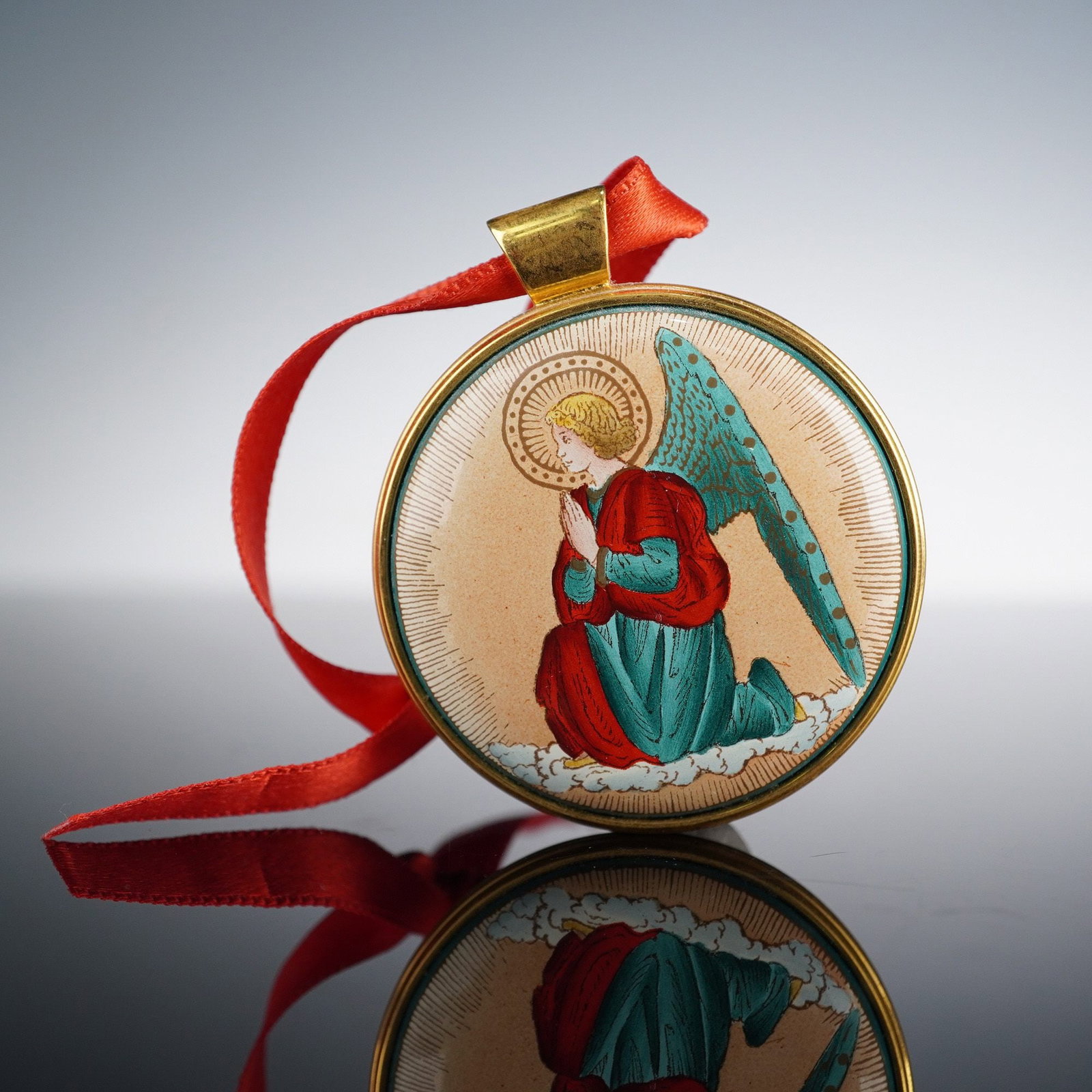 Halcyon Days Enamels for Neiman Marcus Christmas 2000 Kneeling Angel: This circular, gold-tone metal medallion ornament features an enamel or painted depiction of a kneeling, haloed angel with clasped hands and large blue wings on the front, rendered in a traditional st