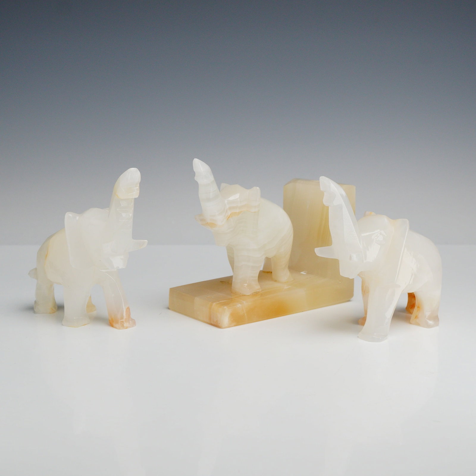 3pc Carved Stone Elephant Figurines including One Bookend: This lot consists of three carved stone elephant figurines, including two freestanding figures and one single bookend. The pieces are crafted from banded onyx, featuring a translucent white and cream 