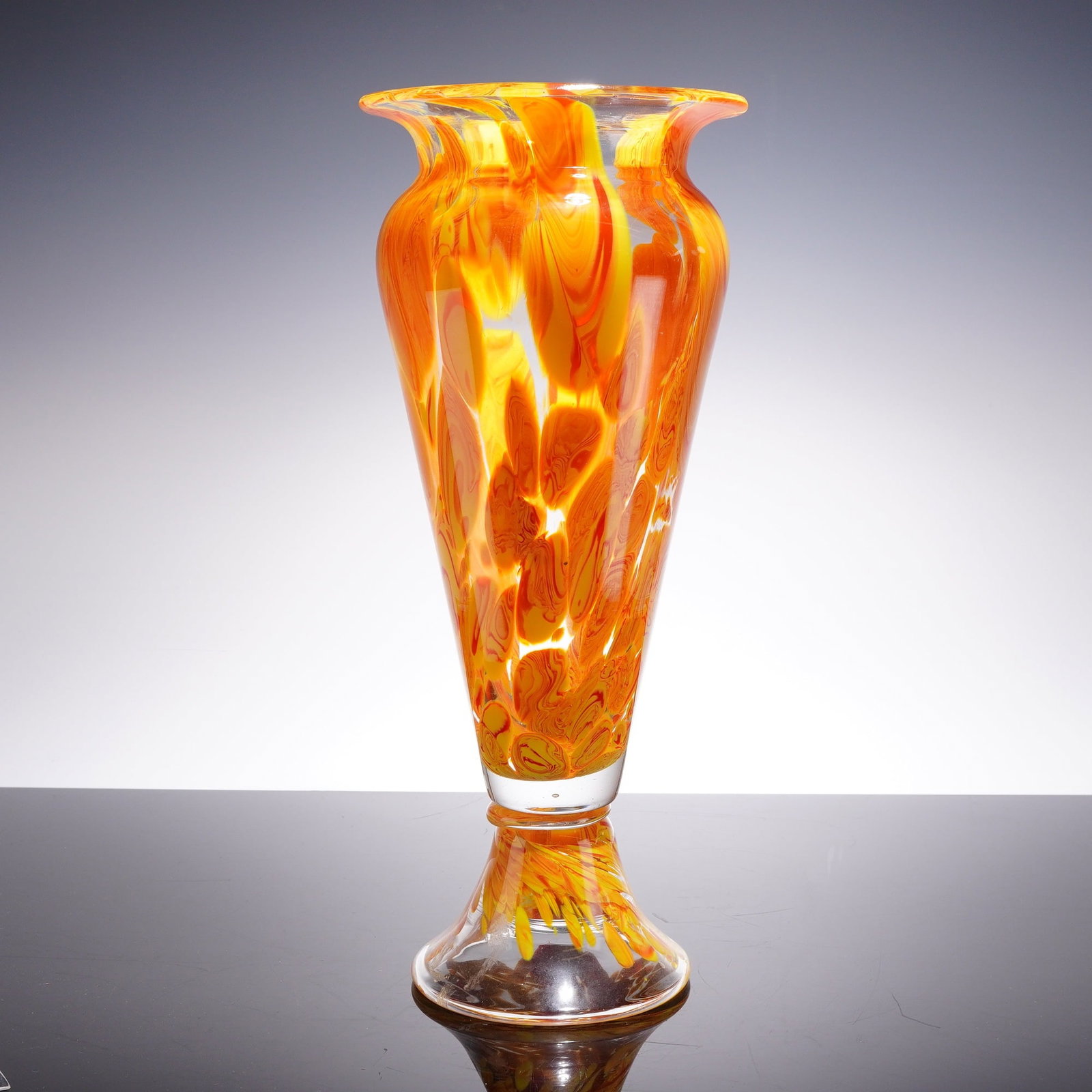 Orange Red and Yellow Hand-Blown Art Glass Vase: This hand-blown art glass vase features a tall, tapered, and elongated form with a bulbous shoulder and a widely flared, slightly flattened rim. The vessel is supported by a clear glass pedestal foot 